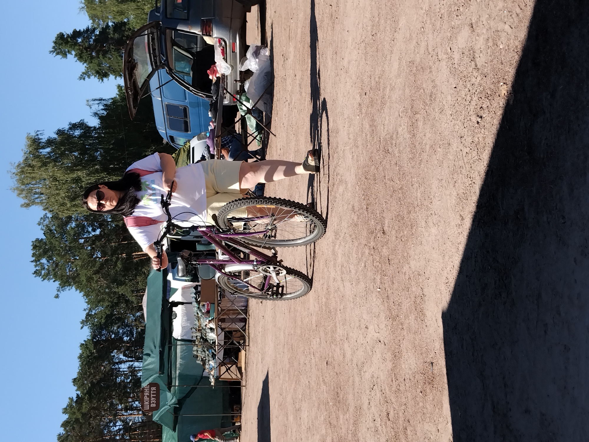 This is a full-body, eye-level, outdoor, medium shot photograph of a woman standing with a bicycle. The woman, who has dark hair and is wearing sunglasses, a white t-shirt, and light-colored shorts, is holding the handlebars of a purple bicycle. The bicycle is positioned in front of her, slightly to her right. She is wearing dark sandals. The woman is standing on a dirt surface. In the background, there are green tents and tables displaying goods, suggesting an outdoor market. Behind the tents, there are trees and a blue van with its rear door open, and a grey car. A person in a red shirt is visible on the left side of the image, walking away from the camera. The sky is clear and blue. The scene appears to be an outdoor market in Irshansk, Ukraine.