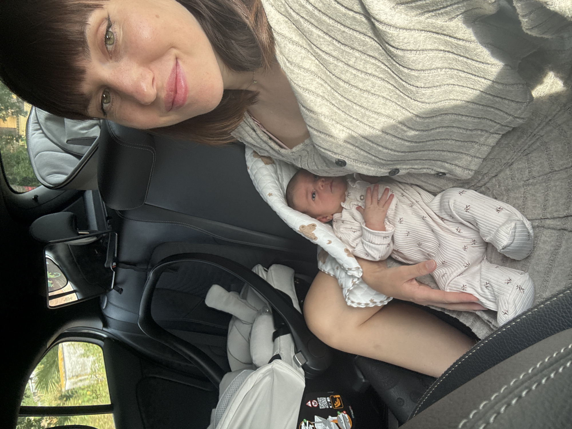 The image shows a woman holding a baby inside a car. The woman has dark hair and is wearing a striped sweater. The baby is wrapped in a blanket. The car's interior is visible, including the car seat. The location is Nettuno, Italy, as indicated by the metadata (not visible in the image itself).