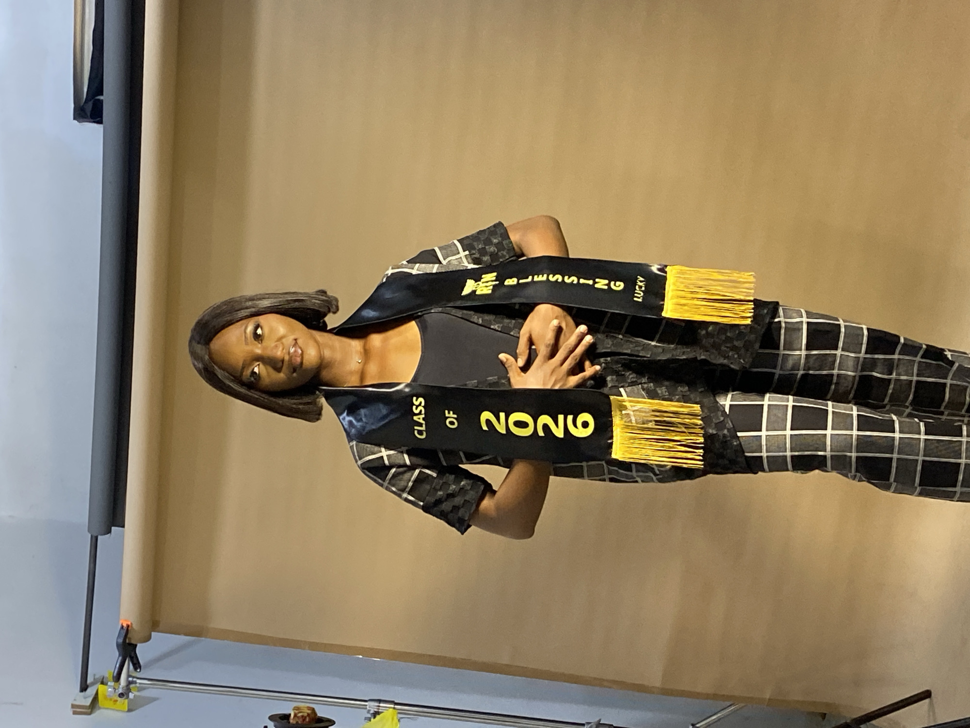 A young woman poses indoors in front of a tan backdrop, presumably at a studio or event space, as suggested by the backdrop support and clamps at the left. She is wearing a black graduation sash that reads "CLASS OF 2026" on one side and "BLESSING LUCKY" on the other, with yellow fringe on both ends. She's wearing a black top underneath a black and white plaid garment. Her hands are interlocked and placed at her center. The specific location in Gura Top, Nigeria cannot be determined from the image.
