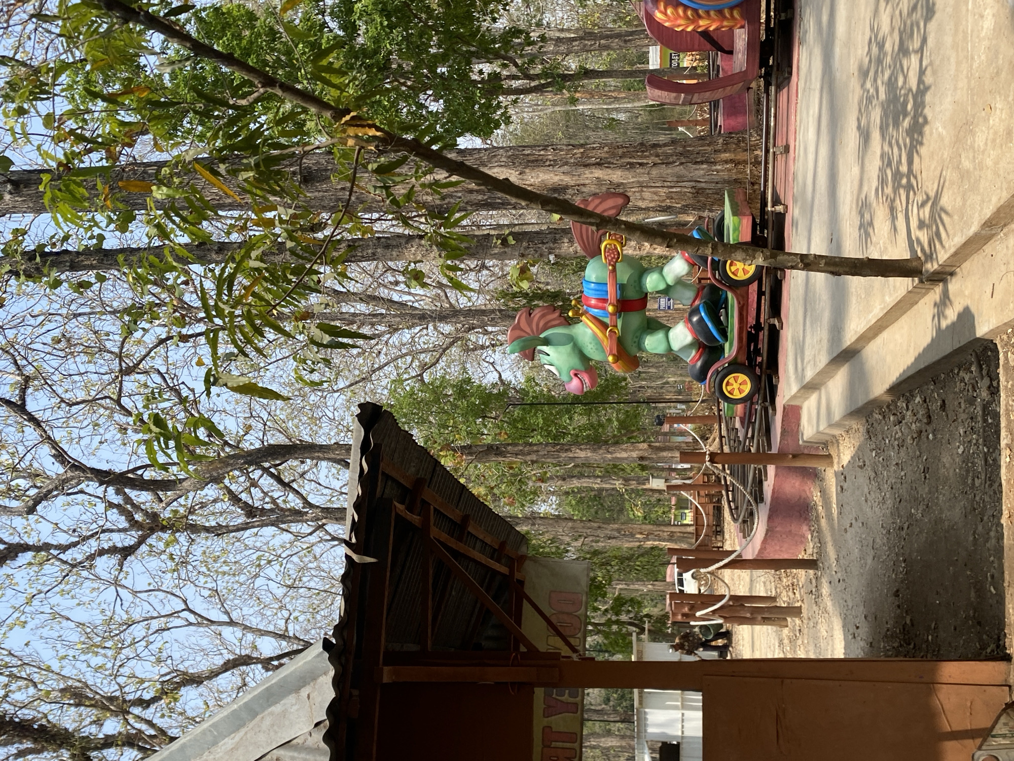 An outdoor amusement park scene is depicted in the image. In the foreground, a light brown wooden structure with a corrugated metal roof is visible on the left. A bright green, cartoonish horse-shaped ride with red and blue accents is positioned on a small amusement park train track. The horse ride is on a green base with yellow wheels. Behind the horse ride, more train cars, one with red and gold decorative elements, are visible on the track. The track is bordered by a low, pinkish-red wall. Wooden posts with white rope are along the track. The background is filled with tall, bare trees with some green foliage. The ground consists of dirt, gravel, and paved areas. Two people are visible in the background, one near a white structure and another near a white vehicle. A sign with the word "PARKING" is visible on the horse ride. The lighting suggests it is daytime with a clear sky.