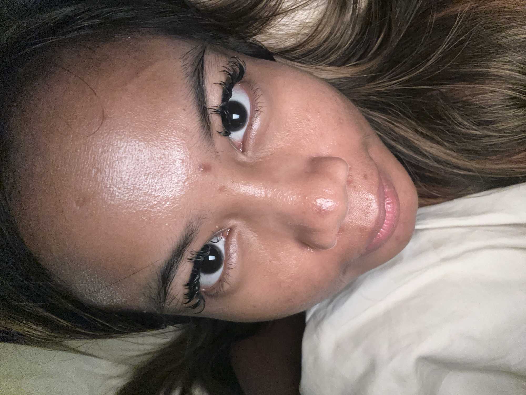 A close-up, tilted view of a woman's face. She has brown hair with blonde highlights. The woman has dark eyes and long eyelashes. Her skin appears somewhat oily. She is lying down on a white sheet. There is no evidence suggesting this image was taken in Ubud District, Indonesia.