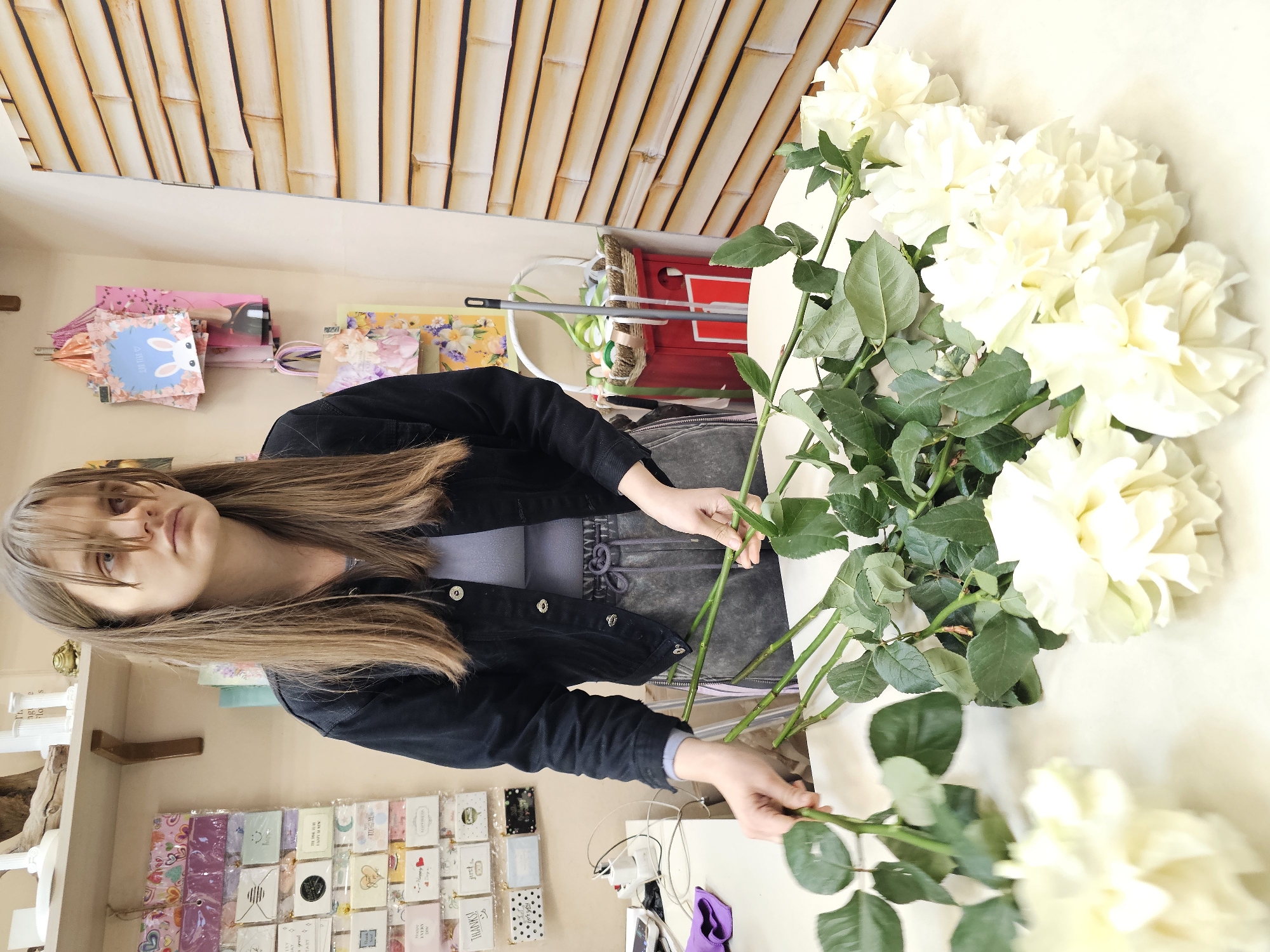 The image, which appears to be rotated 90 degrees counter-clockwise, depicts a young woman in an indoor setting, likely a flower or gift shop in Khimki, Russia. She has long, straight brown hair with bangs and is dressed in a black denim jacket over a grey top. Her hands are actively engaged in arranging or preparing several large, white, fully bloomed roses with green leaves and thick stems, which are laid out on a white table.
Her expression is neutral and focused on her task. In the background, shelves are visible, holding a variety of colorful gift bags, including a pink one and a blue bag adorned with a white bunny, pink cherry blossoms, and partially visible text that appears to say "WITH LUV." Below the woman, numerous small greeting cards are neatly displayed in rows, with legible text like "THANK YOU," "Happy Birthday," "BEST WISHES," and "LOVE," alongside various decorative designs.
The upper background features a distinctive bamboo-patterned wallpaper or paneling. Other items seen include a white power adapter, a smartphone, and a purple piece of fabric. The scene suggests a busy and organized retail or workshop environment, illuminated by natural or artificial daytime lighting.