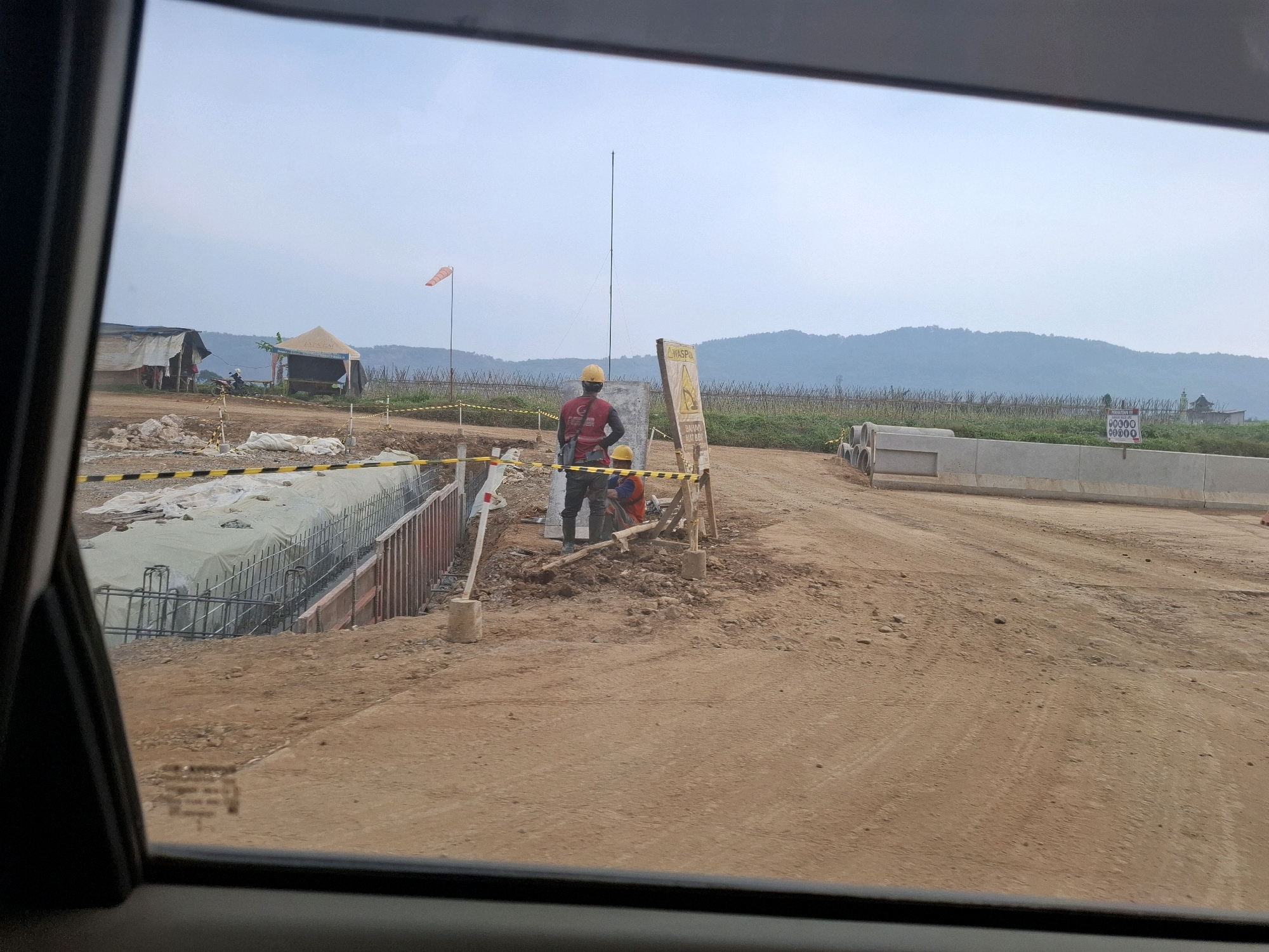 The image captures a construction site in Kutasirna, Indonesia, viewed from inside a vehicle, likely through the windshield. The scene is dominated by a dirt road and ongoing construction work. Two construction workers are visible, both wearing yellow hard hats. One worker, in a red shirt and dark pants, stands near a sign. The other worker, in an orange vest, is kneeling further down the construction area.
In the background, there are temporary structures, including a tent with signage, and what appear to be makeshift shelters. A windsock flutters on a pole, indicating the weather conditions. The construction area itself features excavated trenches with rebar and formwork, suggesting the early stages of building infrastructure, possibly a road or a retaining wall. Concrete barriers and pipes are also present on the site. Beyond the construction area, a gently rolling landscape with hills covered in vegetation stretches towards the horizon under a pale sky. The overall impression is one of an active construction zone in a rural or semi-rural setting within Kutasirna.