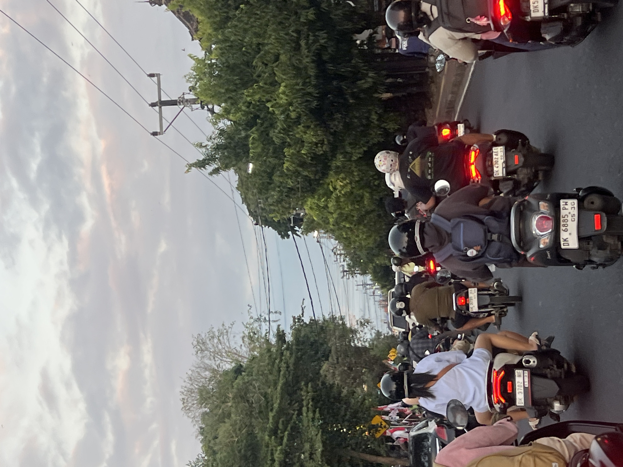 This is a photograph of a traffic jam in Denpasar, Indonesia, as indicated by the license plates on the motorbikes (DK 3702 NE, DK 4702 AEI, DK 5... and DK 6885 PW). The image shows multiple motorbikes with riders, predominantly wearing helmets, navigating a street lined with lush green trees. The sky is overcast with a mix of gray and pinkish clouds. Power lines and poles are visible above the trees. The perspective is from behind a motorbike, looking forward into the traffic. The riders are mostly facing away from the camera, with some looking towards the front and others slightly to the side. The atmosphere appears to be late afternoon or early evening, given the lighting conditions.