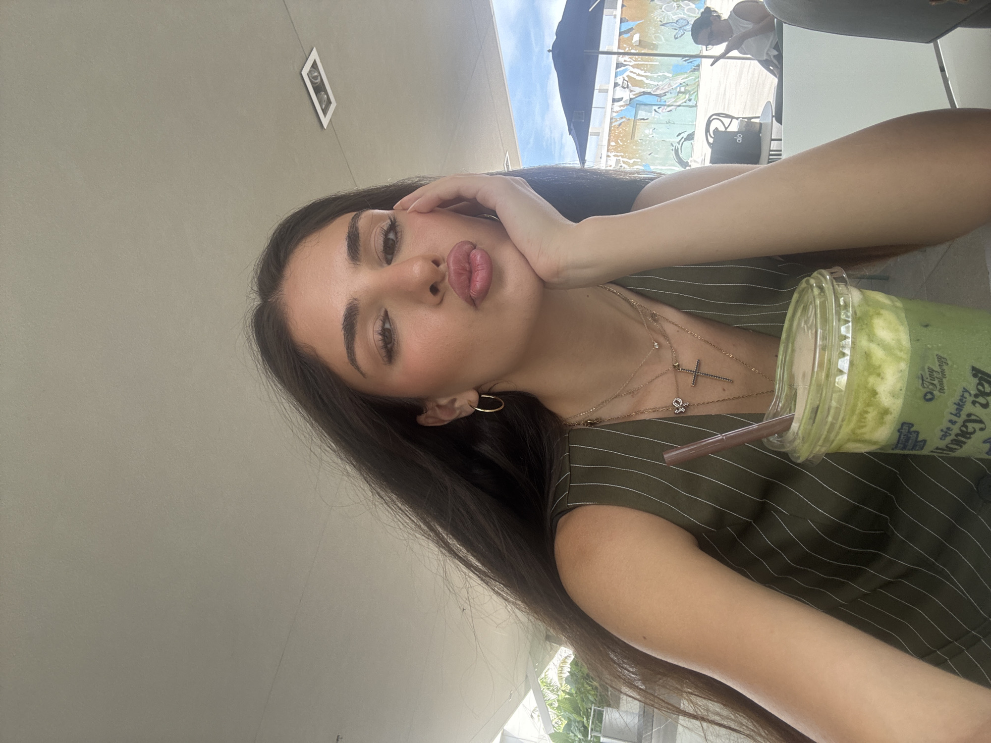 A young woman with long, dark hair, wearing a green pinstripe vest and multiple necklaces, poses for a selfie with her hand on her cheek and her lips puckered. She is holding a green beverage in a clear plastic cup with a straw. In the background, there is a wall with colorful graffiti, an umbrella, and other patrons sitting at tables. The scene appears to be an outdoor seating area of a cafe or restaurant in Hallandale Beach, Florida, USA, under a covered patio with a visible sky.