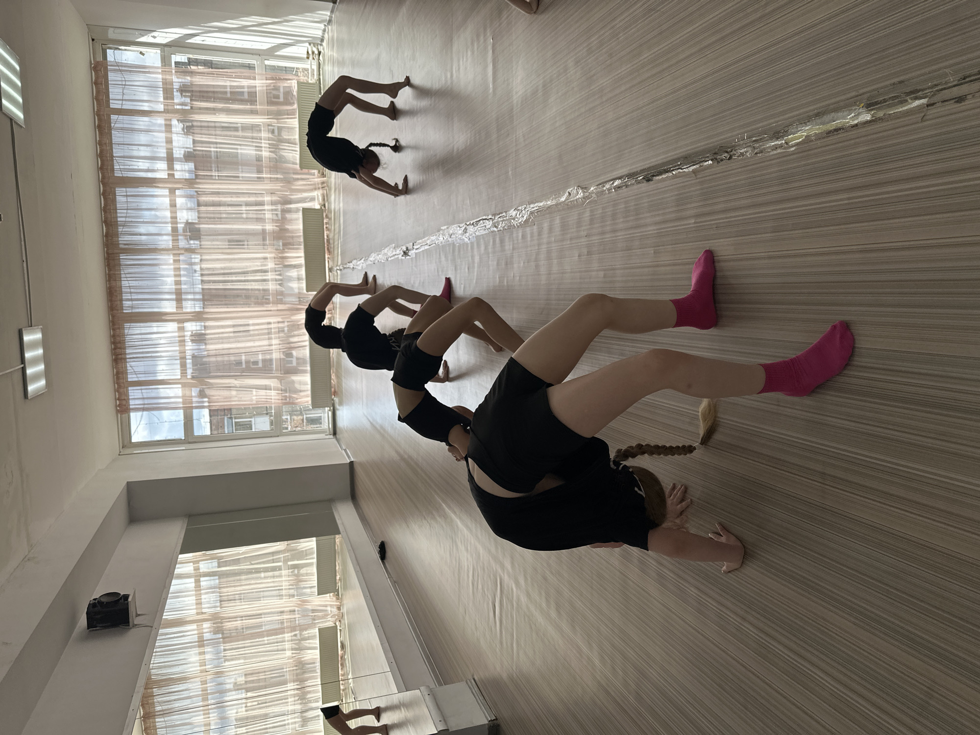 A photo taken indoors, likely in a studio or dance space. The room has large windows covered with transparent curtains. At least three people are performing gymnastic backbend exercises on the floor, arranged in a line. The people are wearing dark-colored clothing. One person is wearing pink socks. The floor is made of light-colored wooden panels. The location is likely a dance studio in Votkinsk, Russia.