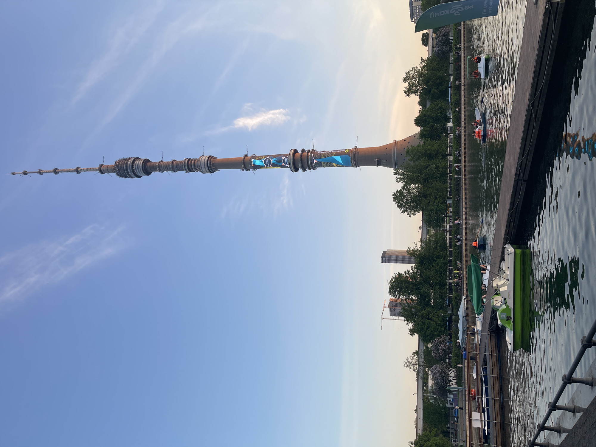 The image depicts the Ostankino Tower in Moscow, Russia, viewed from a lower vantage point that includes a body of water (likely a pond or lake). The tower is the main subject, stretching vertically into a partly cloudy blue sky. The tower is a prominent landmark, with its distinctive segmented design visible. On the water, there are several boats, some with occupants wearing life vests. The banks have greenery with trees. A building under construction is visible in the background, along with other structures. A boardwalk/pier sits on the water. The image is taken during daylight, with diffused lighting suggesting late afternoon or early evening.