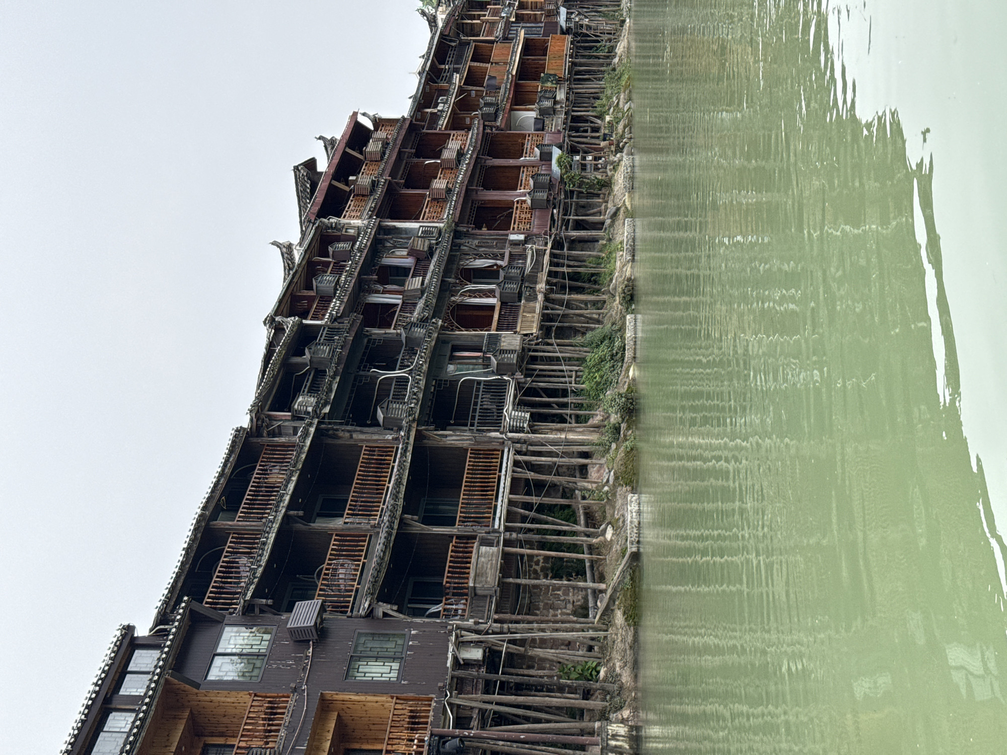 A tall, weathered building, reminiscent of traditional Chinese architecture, stands alongside a body of water. The building features multiple levels with exposed wooden frameworks, balconies, and visible interior spaces. Its facade is aged and appears to be in a state of partial disrepair. The water's surface reflects a distorted image of the building, creating a mirror effect. The environment suggests a location in Fenghuang, China, where such structures are common along the Tuojiang River. The sky above is overcast, casting a muted light on the scene. There are no people visible in the image.