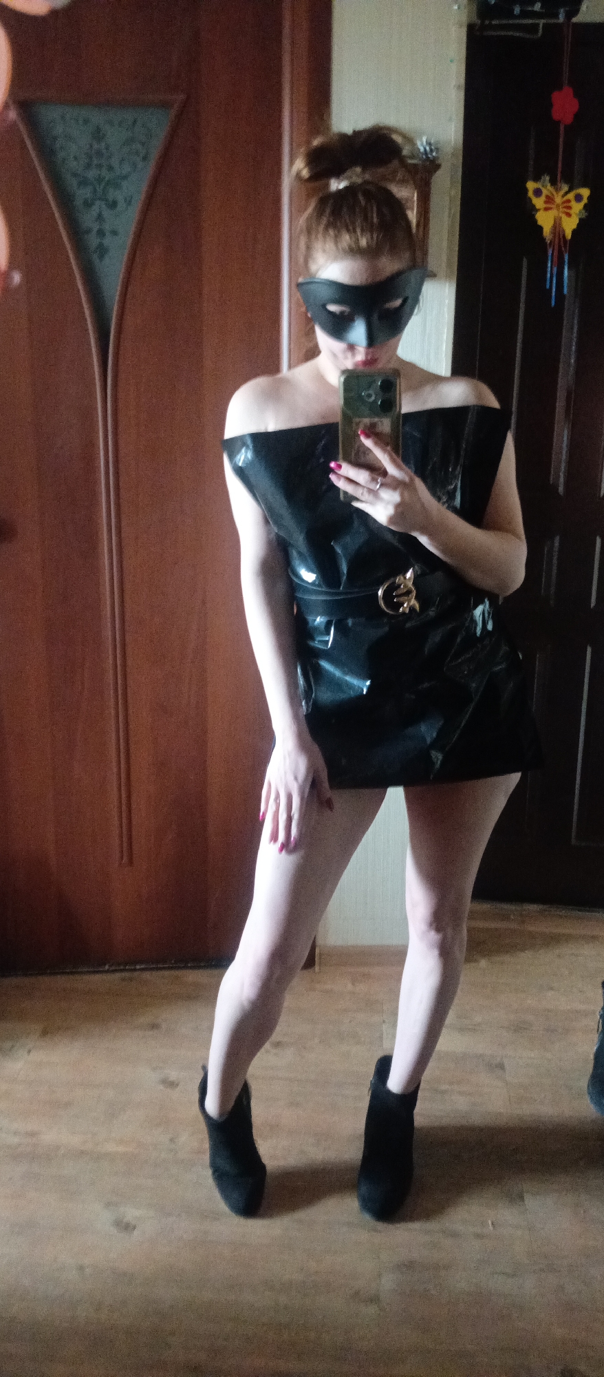 A woman is captured in a mirror selfie, posing indoors. She stands in a slightly dynamic stance, with one leg bent and one hand resting on her thigh. She has reddish-brown hair tied up on her head and wears a black mask covering her eyes and upper nose. Her lips, visible below the mask, are painted red.
Her attire consists of a striking off-the-shoulder black dress or top, which appears to be made of shiny material like latex or vinyl, cinched at the waist by a black belt featuring a prominent gold-colored, possibly intertwining, buckle. The dress is short, revealing her bare legs, and she completes her look with black ankle boots. Her fingernails are painted red, and she wears a ring on the ring finger of her left hand. She is holding a smartphone, likely an iPhone with multiple cameras, in her right hand to take the photograph.
The background shows an interior setting. To the left, there's a wooden door with a decorative glass panel. To the right, another dark wooden door is visible, next to which hangs a decorative item resembling a yellow butterfly with blue and red tassels. The floor is made of light-colored wood or laminate. No text is visible in the image.