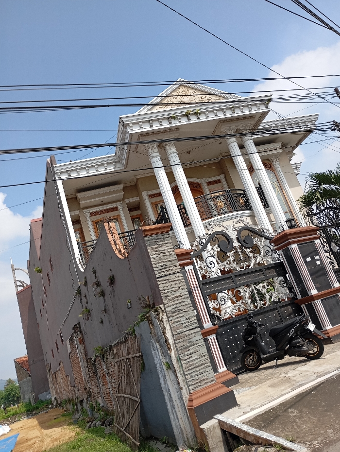 The image shows a two-story house with a white facade and ornate details, situated in Kutawaringin, Indonesia, at coordinates -6.978, 107.549. The house has a balcony with a wrought iron railing and several large columns. The house is partially obscured by a tall, gray wall with a wooden fence and a gate with a black wrought iron design. A black motorcycle is parked on the ground in front of the gate. There are numerous electrical wires running across the sky, and a clear blue sky above. The image is taken from a low angle, looking up at the house and the wall.