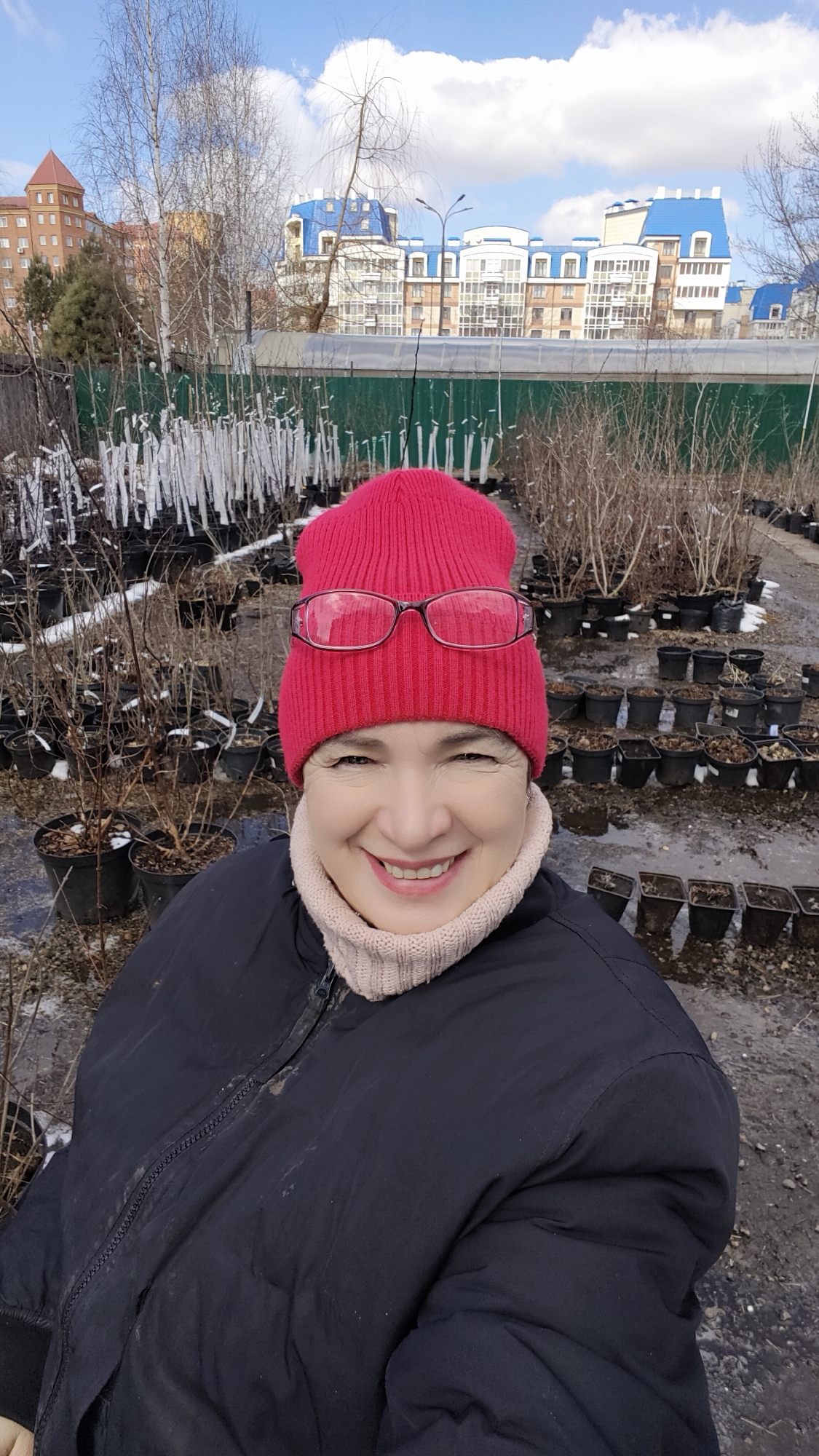 A middle-aged woman with fair skin is smiling at the camera while wearing a pink beanie and glasses. She is dressed in a black winter jacket with a beige turtleneck sweater underneath. The backdrop consists of rows of potted plants, most of which appear to be dormant or bare, with some wrapped in white labels. Behind the plants, there's a green fence and a view of buildings with distinctive blue roofs. The sky above is partly cloudy. The ground is a mix of soil and paved areas, suggesting the setting is a plant nursery or garden center within an urban environment.
