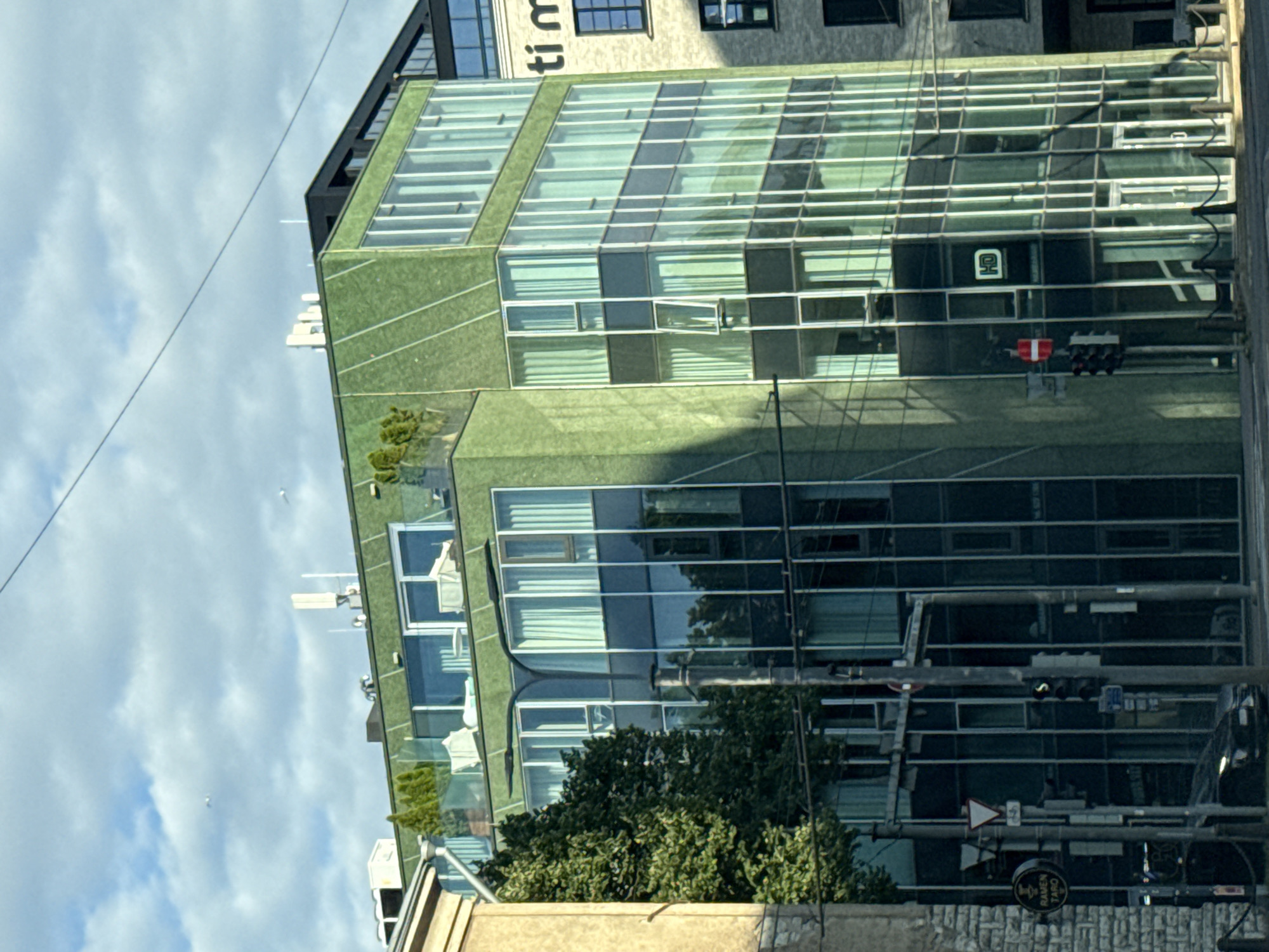 The image shows a modern, multi-story green building with extensive glass facades. The building is located in Tallinn, Estonia, specifically at the intersection of Narva maantee and Laikmaa tänav, as indicated by the street signs and the architectural style. The building features a rooftop terrace with greenery and outdoor furniture. On the lower floors, there are multiple windows, some of which are open. A traffic light is visible on the left, with a red signal illuminated. There are several street lamps and electrical wires present. A tree with dense foliage stands to the left of the building. In the background, another building with "ti m" visible on its facade is partially obscured. The sky is partly cloudy. There are no people visible in the frame. The overall scene depicts an urban environment with contemporary architecture.
