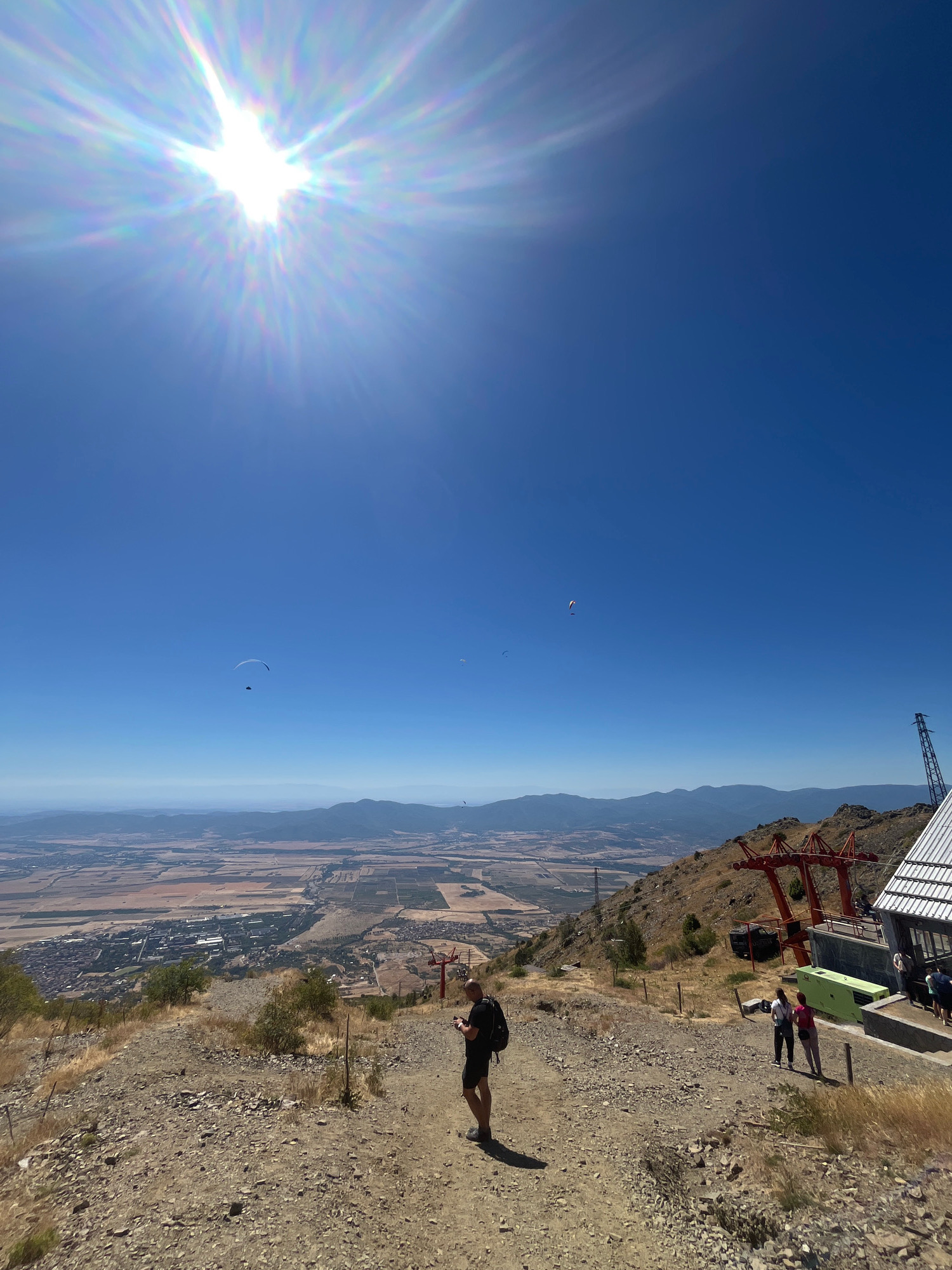 The image depicts a high-altitude view overlooking the city of Sopot, Bulgaria. In the foreground, a dirt path leads down a rocky, sparsely vegetated slope. A lone individual wearing a backpack stands on the path, facing away from the camera. Further down the slope, near a ski lift station, two women in casual attire are visible, one with a white backpack, and another in a pink top. A ski lift structure, painted red, is prominent, along with a modern building and a green utility box. In the sky above, several paragliders are seen suspended against a clear blue sky. The sun, a bright, starburst of light with visible lens flares, dominates the upper left quadrant of the image. The background reveals a valley with agricultural fields, the town of Sopot with its buildings, and a range of distant, hazy mountains under a bright, sunny sky. The overall scene suggests an outdoor recreational area, likely a popular spot for paragliding and hiking.