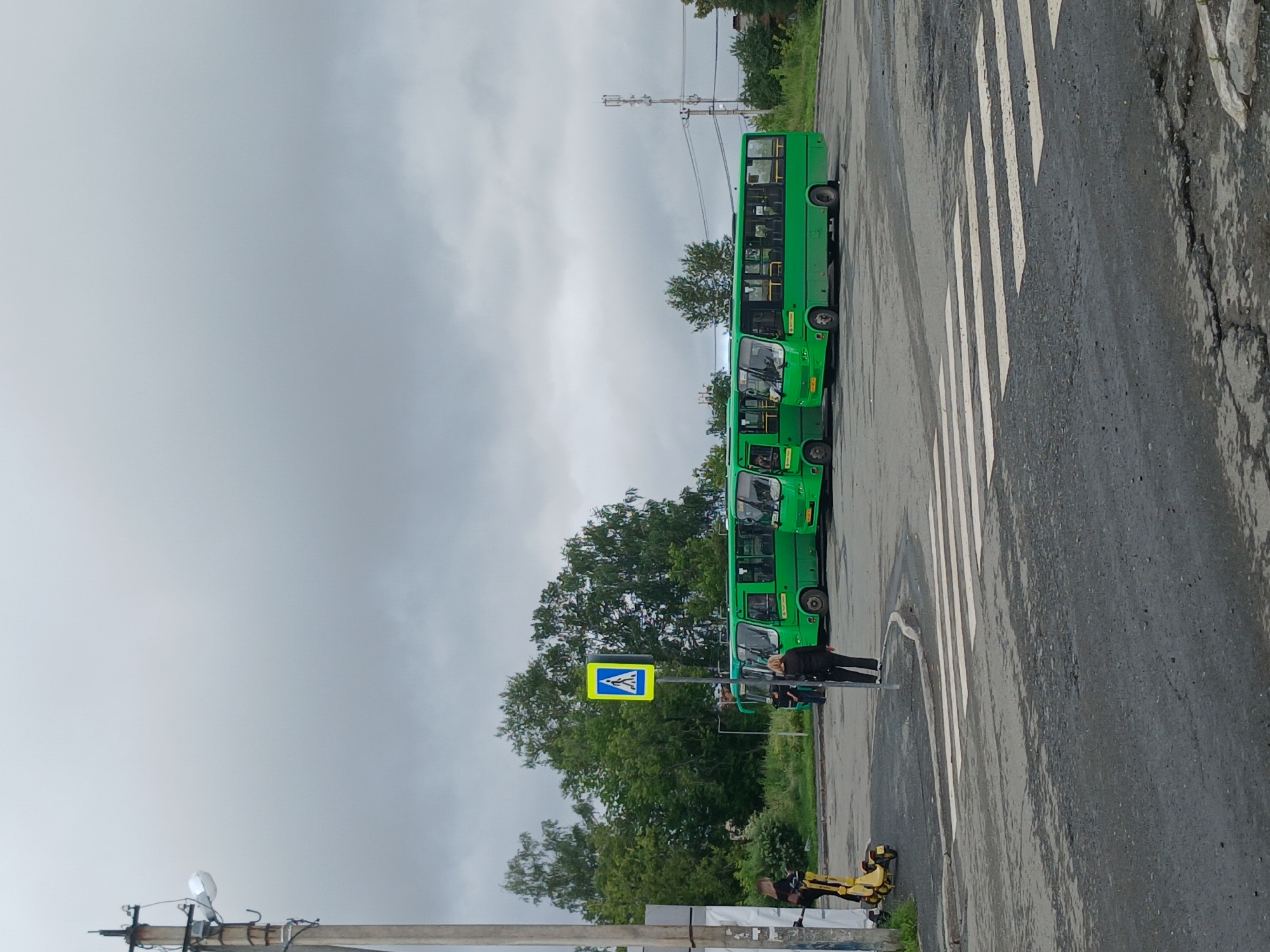 In Yekaterinburg, Russia, on a cloudy day, a street scene unfolds featuring several green buses parked in a lot. A pedestrian crossing with white stripes is visible on the asphalt in the foreground. To the left, a woman stands next to a yellow scooter. Further into the scene, a pedestrian crossing sign with a yellow background and a black silhouette of a person walking is mounted on a pole. Three green buses are parked in the background. The sky is overcast, and trees are visible behind the buses. A utility pole with wires and a light fixture is present on the far left. Another utility pole with antennae is visible in the distance on the right.