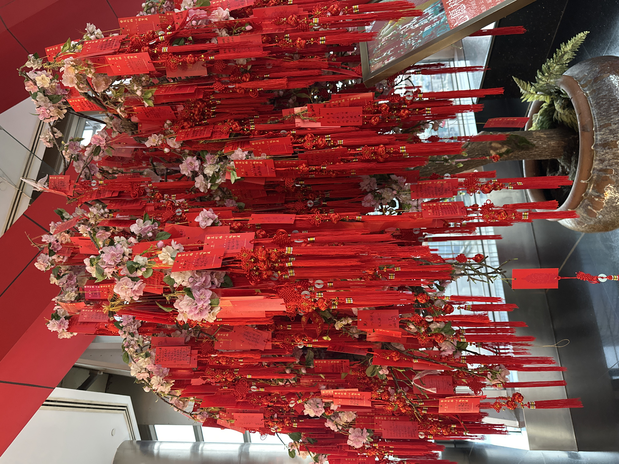An artificial tree, adorned with numerous red tags and tassels, is displayed in a large ceramic pot. The tree has pink and white artificial flowers. The red tags, which appear to have handwritten inscriptions, are attached to the branches along with decorative red knots and small golden accents. Behind the tree, a window reveals a cityscape, suggesting an indoor setting with a view of Pudong, China. A framed poster is partially visible to the right of the tree. The scene is illuminated with bright, artificial lighting. There are no people visible in the image.