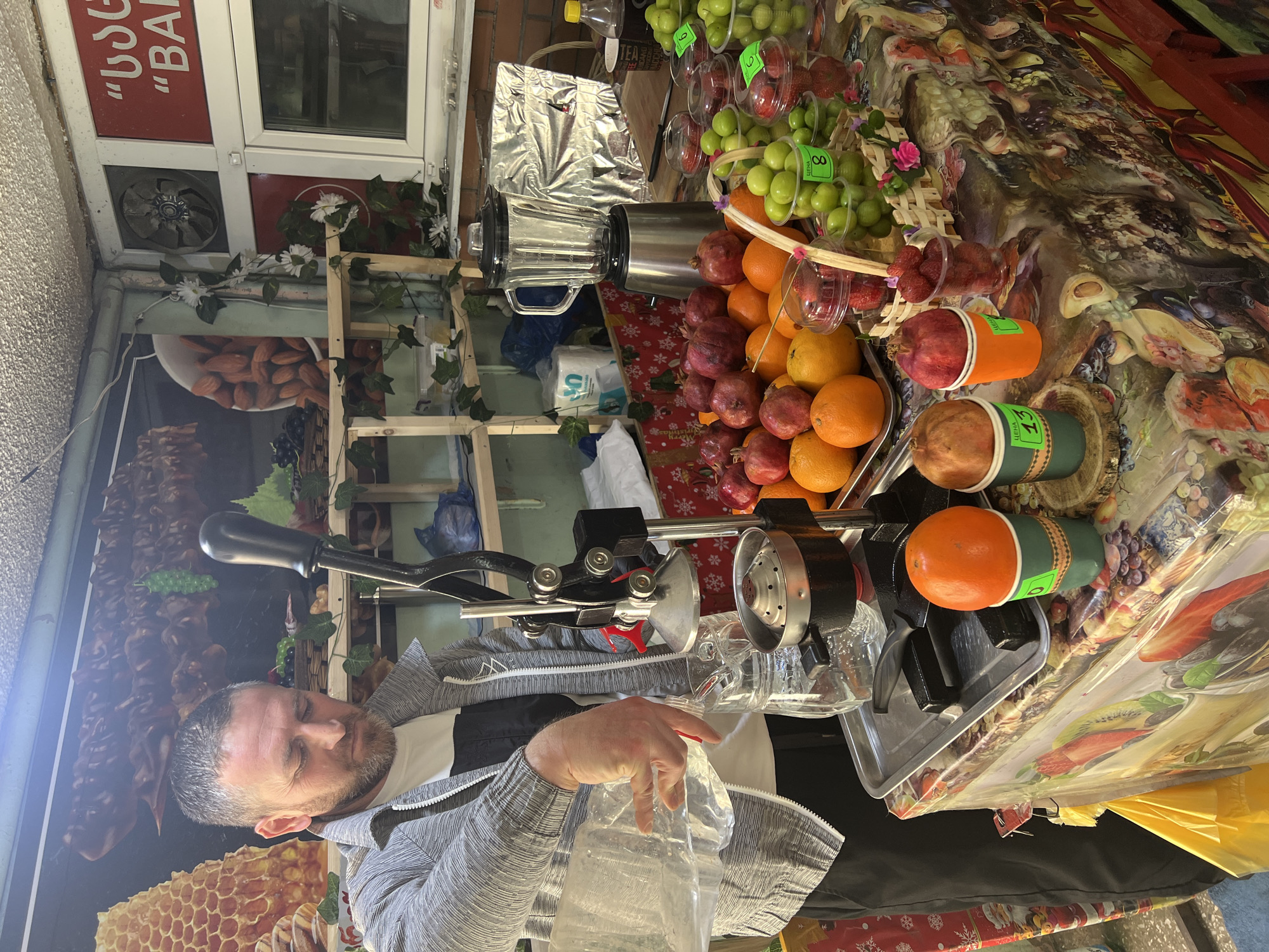 A man in a grey jacket is standing at a fruit stand in Tbilisi, Georgia. He is holding a clear plastic bag under a metal juice press. The stand is covered with a patterned plastic tablecloth and displays various fruits, including oranges, pomegranates, grapes, and strawberries. The fruits are packaged in clear plastic containers or arranged in baskets. Behind the stand, there is a wooden shelf and a building facade with a sign that reads "BAR" in Georgian and English. A blender and other supplies are visible on the shelves. The lighting is somewhat dim, suggesting an indoor or shaded location.