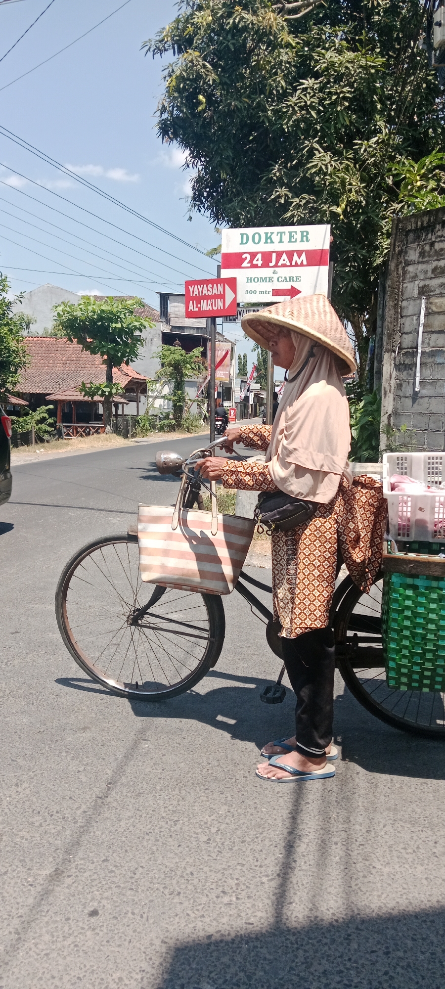 A woman wearing a conical hat and a patterned dress with a headscarf is standing next to a bicycle on a paved road in Maguwoharjo, Indonesia. She is holding the handlebars of the bicycle, which has a large striped bag hanging from it. A green basket is attached to the rear of the bicycle. In the background, there are buildings, trees, and a sign that reads "DOKTER 24 JAM & HOME CARE 300 mtr." The sky is blue with some clouds. The overall scene depicts a street view with a woman and her bicycle, likely a vendor or someone traveling in the area.