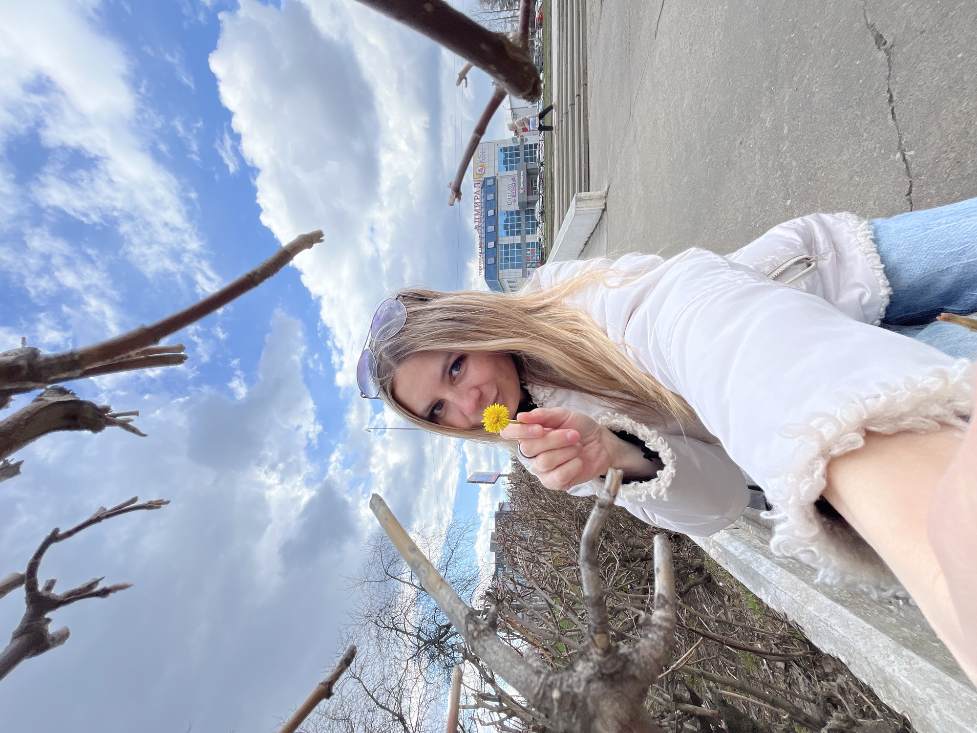 A woman with long blonde hair is holding a dandelion. She is wearing a white jacket and blue jeans. Sunglasses are placed on her head. The photo is taken from a low angle, looking up at her. In the background, there is a cloudy sky, bare tree branches, and a building. The building has "АДМИРАЛ" written on it in Cyrillic letters. Based on the building's name and architectural style, the location is likely Chaikovsky, Russia.