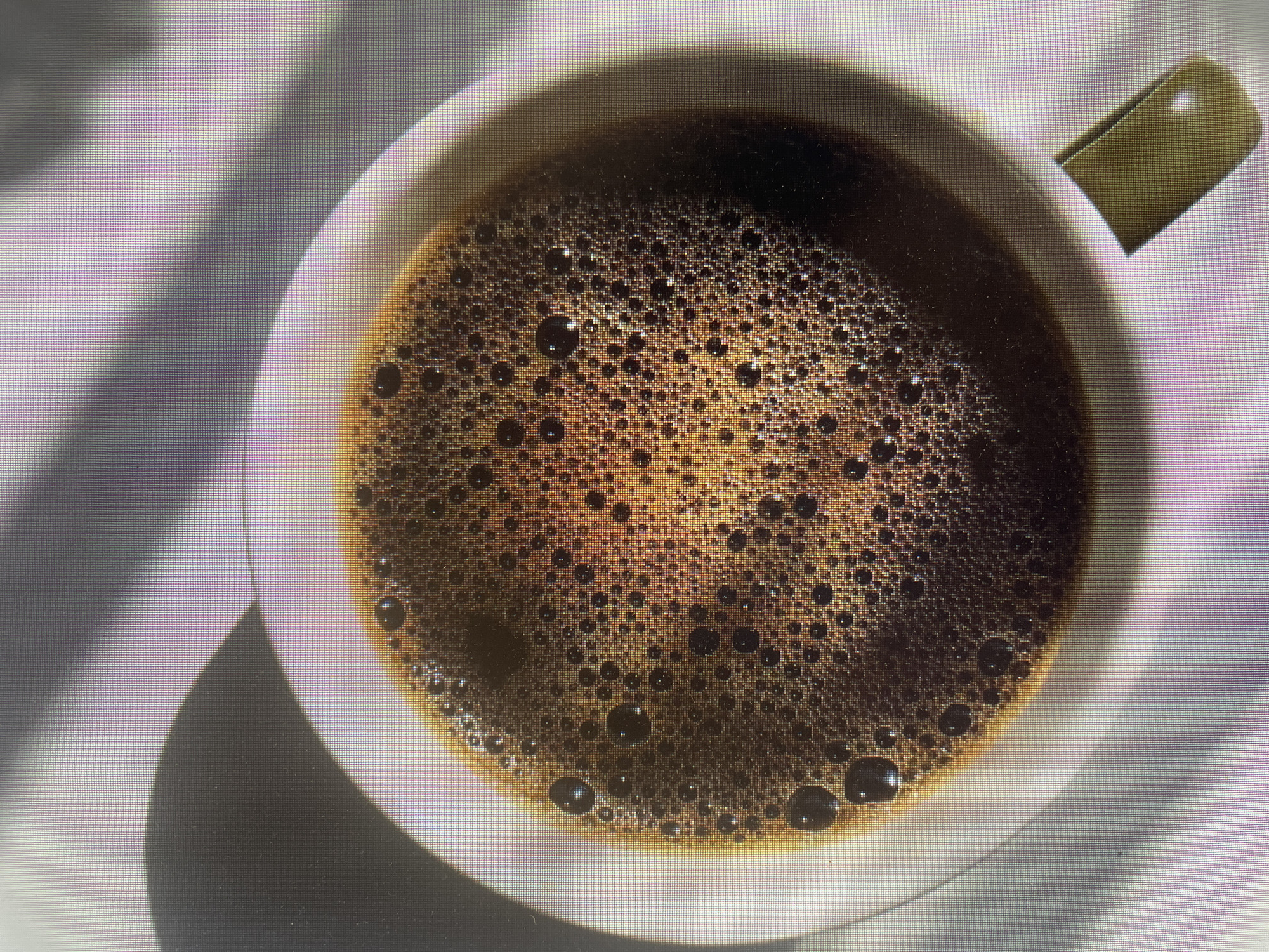 An overhead, close-up shot captures a white or off-white ceramic cup filled with black coffee, set against a light-colored surface. The entire image is overlaid with a prominent, uniform grid-like pixel pattern, suggesting it is a photograph of a digital screen.
The coffee inside the cup is a rich, dark brown, topped with a thick layer of fine bubbles and crema of varying sizes, indicating it is freshly prepared. An olive-green handle is partially visible on the right side of the cup.
Strong, directional light, likely from the upper left, casts distinct shadows. A large, softly diffused shadow extends from the cup on the left, while the handle creates a sharper, more defined shadow on the right. The contrast between light and shadow highlights the contours of the cup and its contents. The scene implies a moment of quiet contemplation or a beverage break. No people or text are visible in the image. The setting appears to be indoors, possibly in Poltava, Ukraine.