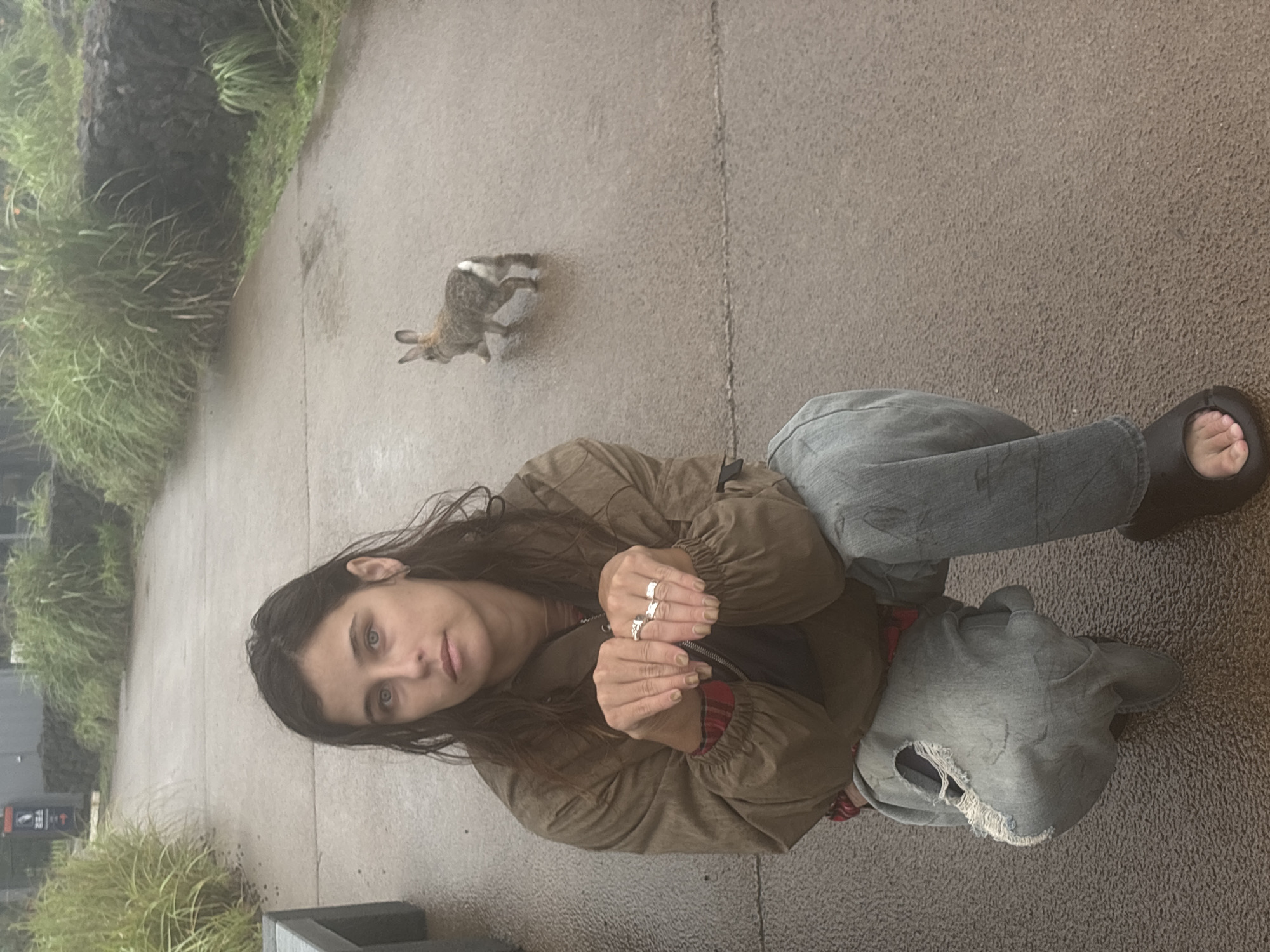 The image features a young woman with long, dark hair and striking blue eyes, crouching on a paved surface. She is wearing a brown jacket over a red and black plaid shirt, and loose-fitting denim jeans. Her hands are clasped in front of her, adorned with several rings. To her left, a small, furry rabbit is captured mid-hop on the same paved ground. Lush green vegetation lines the left side of the frame, suggesting an outdoor setting. The pavement appears damp, hinting at recent rain or mist, and the overall lighting is diffused, indicating an overcast day. The woman's expression is calm and observant, her gaze directed towards the viewer. Beside her, a grey, ripped hooded sweatshirt rests on the ground. The scene is set in Seogwipo-si, South Korea. There is some faint text visible on a sign in the background, but it is illegible.