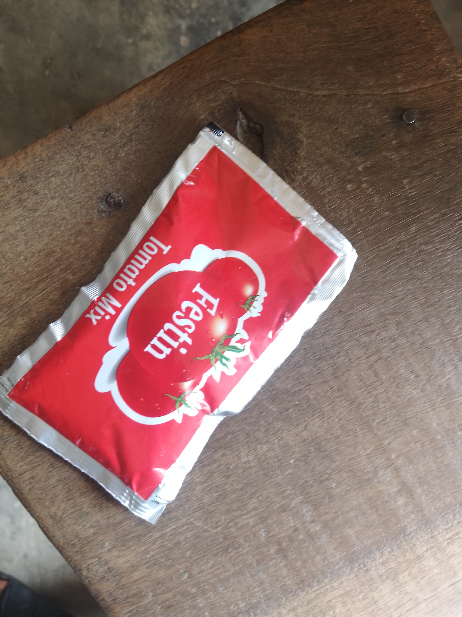 A red and white packet of "Festin Tomato Mix" lies on a rustic wooden surface. The packet is oriented diagonally, with the text "Tomato Mix" written vertically in white on the left side. The brand name "Festin" is displayed prominently in large white letters on a stylized tomato graphic in the center of the packet. Two small green tomato vines with leaves are depicted below the brand name. The wooden surface appears weathered and has visible grain and texture. The overall scene is simple and focuses on the product. This image was captured in the city of Serti, Nigeria.