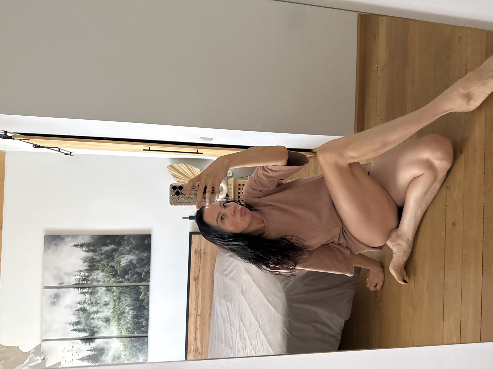 A woman is sitting on a wooden floor in front of a mirror. She is wearing a light pink t-shirt and has long, dark hair. Her legs are crossed, and she is holding a phone in her right hand, taking a selfie. In the background, a bed with a grey comforter is visible, along with a triptych of forest artwork on the wall. A sliding barn door is also present to the right of the woman. The scene appears to be a bedroom. The location is not in Chaikovsky, Russia.
