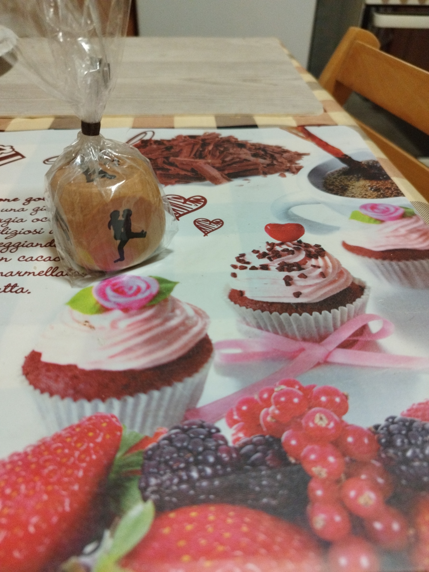 A close-up shot depicts a pastry item in a clear plastic bag, sealed with a dark band around its neck. A silhouette of a couple is printed on the pastry. The pastry sits on a tablecloth printed with images of cupcakes, chocolate shavings, a cup of coffee, and an assortment of berries including strawberries, blackberries, and red currants.
There is no evidence of the location being in Sofia, Bulgaria.