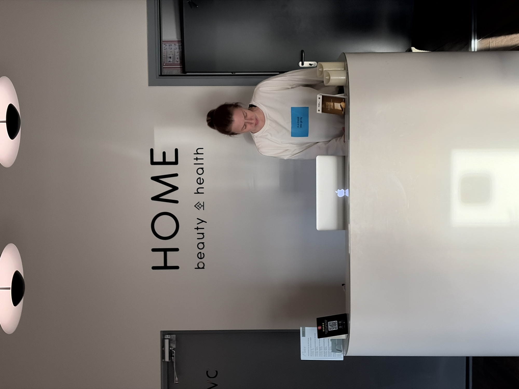 The image shows an indoor reception area, possibly in the city of Бараўлянскі сельскі Савет, Belarus, judging by the presence of Russian language text on signage. A woman with her hair in a bun stands behind a white reception desk, looking down. She is wearing a white sweatshirt with a blue rectangular patch on it. On the desk, there is a laptop with an Apple logo, a small digital display showing a QR code, and two decorative candles. To the left of the desk, there is a door with "VC" written on it, and a sign with text and a QR code. A larger sign with black lettering reads "HOME beauty & health". To the right of the woman is a dark door. The overall impression is a clean, modern reception area.