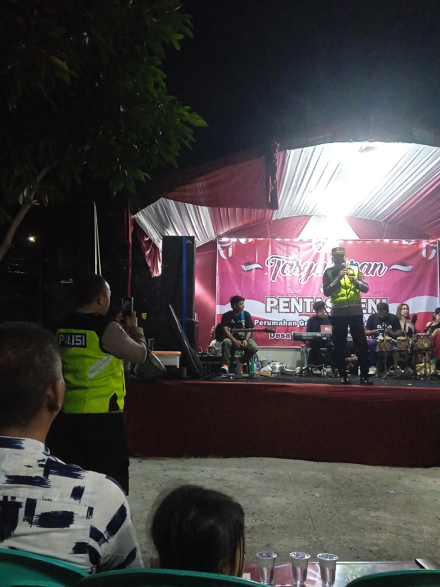 The image depicts an outdoor event at night, possibly in Cangkring, Indonesia. A stage is set up with a pink banner that reads "Tasyakuran PENTAS SENI" and "Perumahan Grand Desa." On the stage, a person in a police uniform with a reflective vest is speaking into a microphone. To the left of the speaker, there is a musical ensemble with several individuals playing instruments, including drums and a keyboard. Several audience members are seated in the foreground, some of whom are wearing casual clothing. A police officer in a reflective vest is standing on the side of the stage, holding a mobile phone, seemingly taking a picture or video. The environment appears to be a gathering or festival, illuminated by stage lighting and ambient darkness.