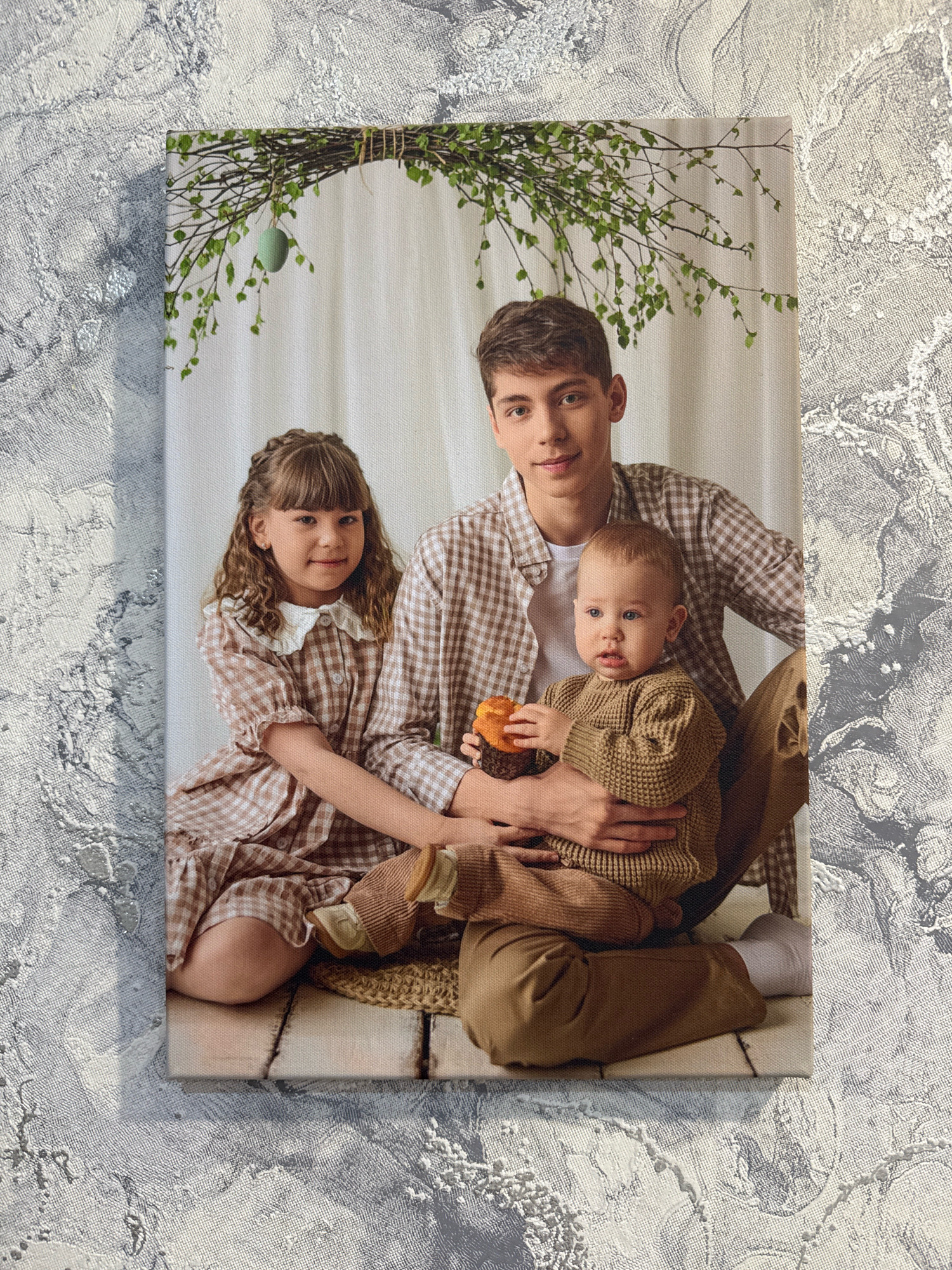 The image displays a canvas print of a family posing for a photograph. Three individuals are visible: a young man, a young girl, and a baby. The young man is standing behind the other two, with his arms around the baby. The young girl is seated in front of the man. All three are dressed in coordinated brown and white checkered outfits. The background consists of a white curtain, with a branch adorned with green leaves and a single green egg hanging overhead. The canvas is placed on a surface with a textured, grey and white pattern. The provided location, Rusanovo, Russia, is not visually depicted in the image itself.
