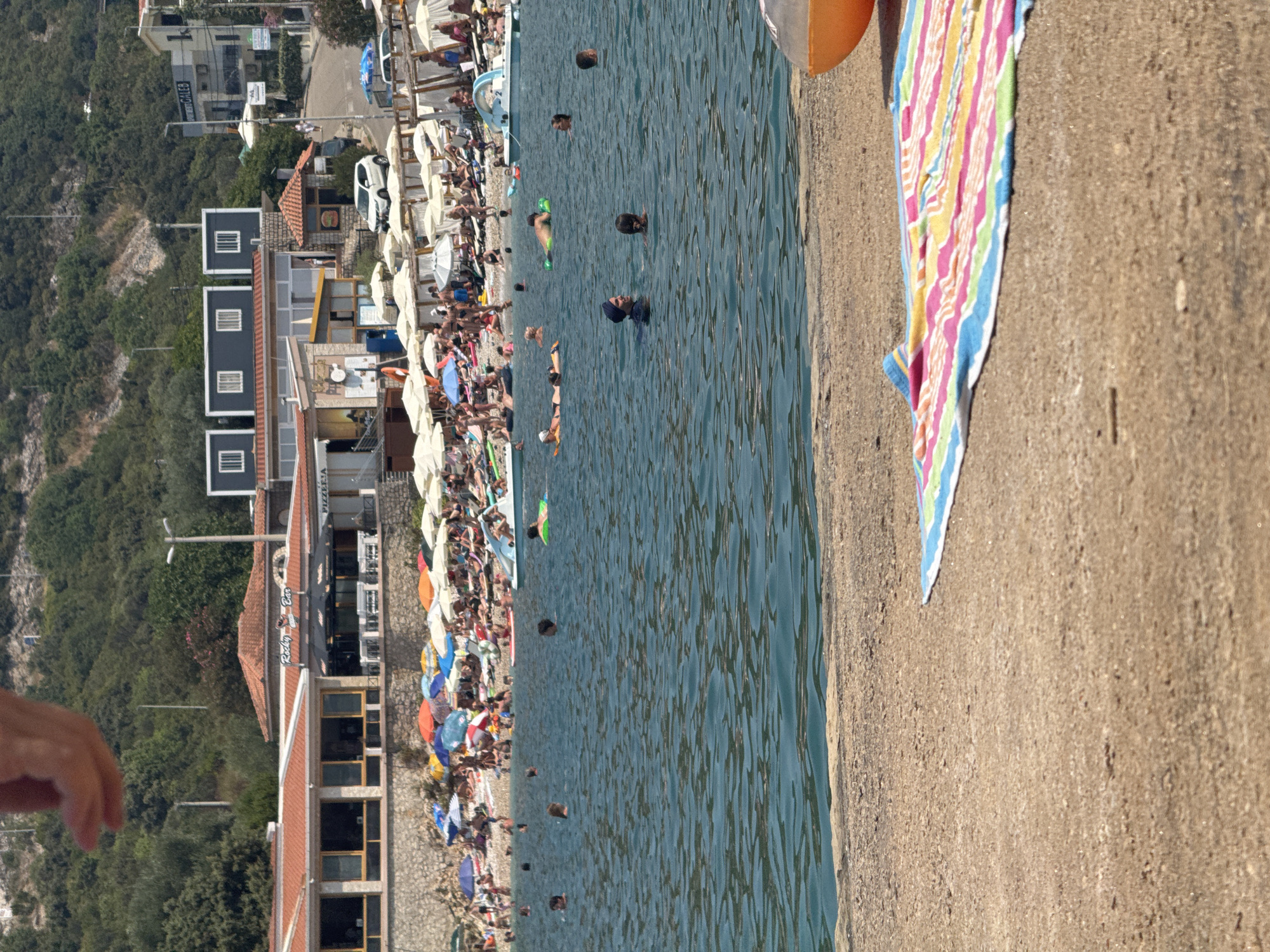 This is a medium shot of a crowded beach on a sunny day in Kunje, Montenegro. The foreground shows a sandy beach with a colorful striped towel laid out. The water in the midground is a bright blue and several people are swimming or floating. In the background, there is a densely packed beach with many people relaxing under umbrellas, sunbathing, and wading in the shallow water. Behind the beach, buildings line the shore, including a restaurant with a "Pizzeria" sign, and further back, a hillside covered in trees rises steeply. There are also several boats docked in the water. The overall impression is one of a lively and popular tourist destination.