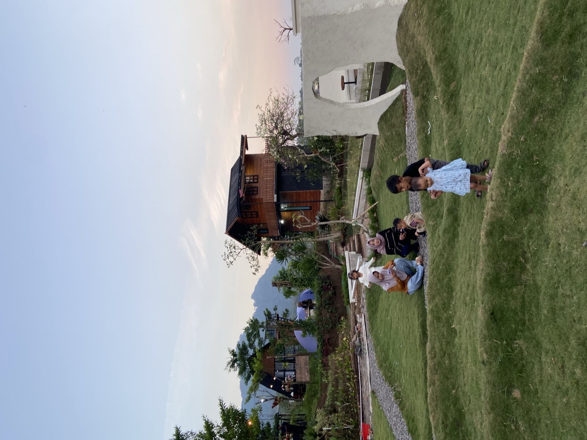 This photograph captures a serene outdoor scene at what appears to be a tourist spot or resort in Kesiman, Indonesia. The setting is characterized by rolling green hills, lush vegetation, and a backdrop of mountains under a pale blue and pink sky, indicating either early morning or late afternoon.
In the foreground, several individuals are enjoying the ambiance. Two women and a young child are seated on the grass. One woman, wearing a yellow hijab and orange top, is looking at her phone. Beside her, another woman in a striped shawl and dark top is looking away from the camera. A young child in a light-colored outfit sits near them. Further to the right, a young boy in a black t-shirt is guiding a toddler dressed in a blue striped dress down a grassy slope.
In the mid-ground, a rustic wooden house with dark windows and a balcony stands prominently, nestled amongst trees. To its left, another A-frame style building with glass walls is visible, alongside a blue tent. The area is landscaped with pathways and planters.
In the background, a majestic mountain range looms under the soft hues of the sky. A large, abstract white sculpture with a curved opening is also a notable feature on the right side of the frame, framing a view of a small table and chairs beyond. The overall impression is one of peaceful recreation in a scenic natural environment.