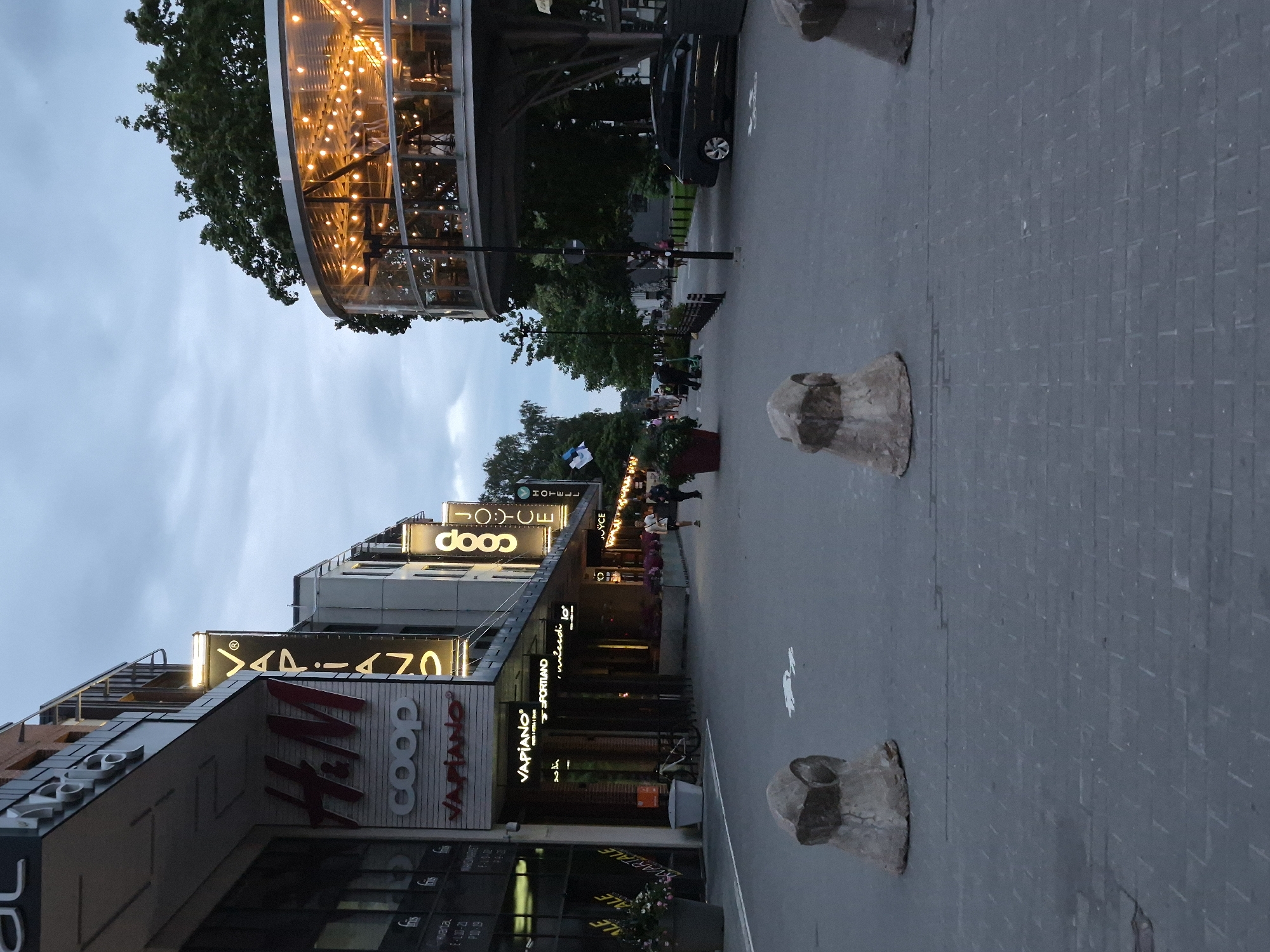 A street scene in Tartu, Estonia, captured in low light conditions under an overcast sky. The foreground features a paved pedestrian area with three concrete bollards, each shaped like a rounded cone with a rough, textured surface. The pavement is made of dark grey interlocking bricks.
In the midground, the street opens up, lined with buildings on the left and trees and a modern, circular, glass-enclosed structure with illuminated interior lights on the right. The buildings on the left display prominent signage for various businesses, including "H&M," "COOP," and "VAPIANO." People are visible walking along the street. To the right, a dark-colored car is parked on the side of the road.
The background is dominated by trees and the sky, which is cloudy and grey, suggesting it is either early morning or evening. The overall atmosphere is subdued, likely due to the twilight conditions. The architecture suggests a modern urban environment. The circular structure on the right appears to be a restaurant or bar, with many warm lights visible within its transparent walls, creating a festive contrast to the surrounding dim light. An Estonian flag is visible on a pole on the left side of the street.