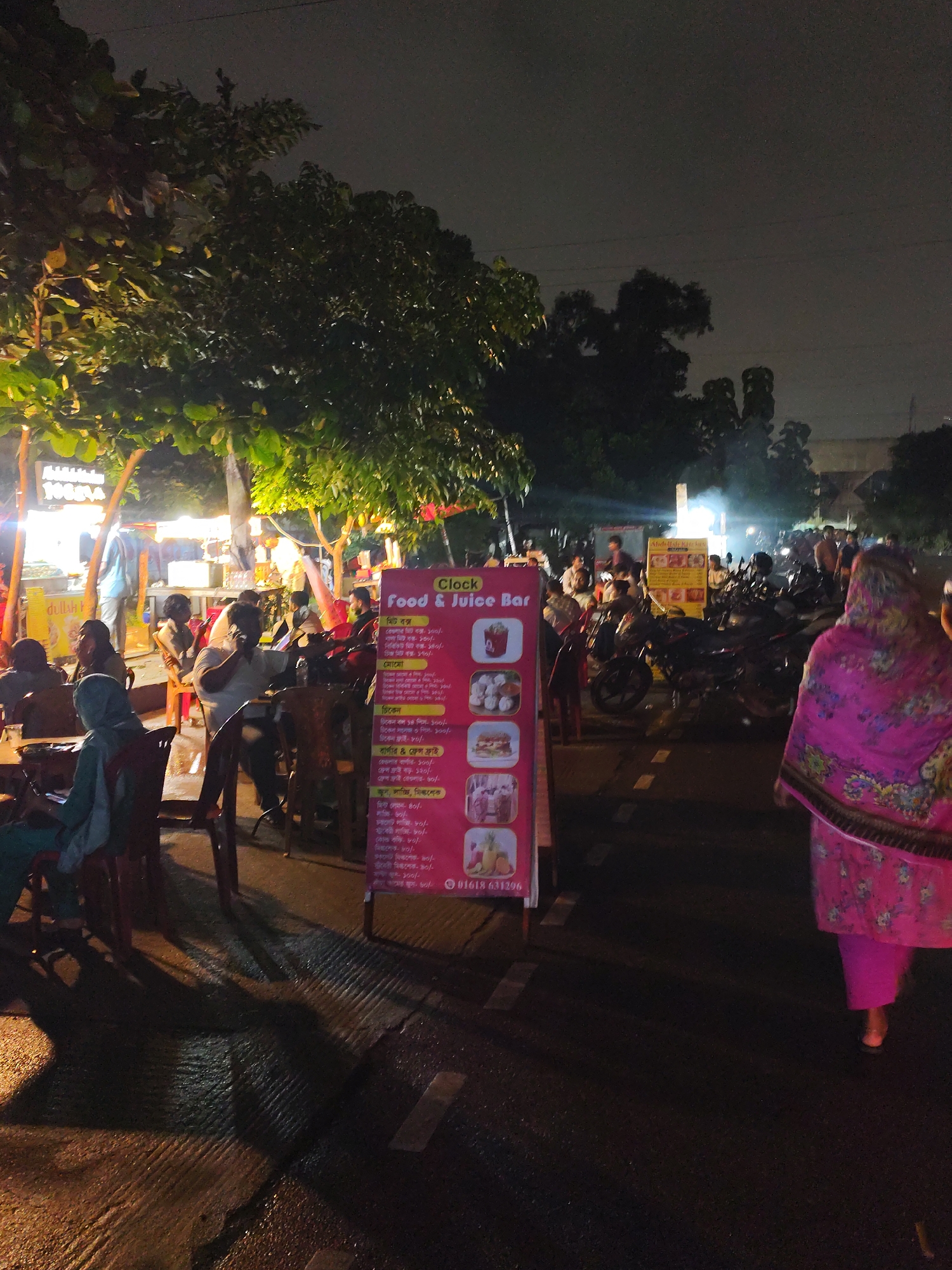 This is a night-time, eye-level, medium shot of an outdoor food and juice bar scene in Dhaka, Bangladesh. In the foreground, a bright pink and floral-patterned shawl is worn by a woman walking away from the camera towards the right. In front of her, a pink sign for "Clock Food & Juice Bar" stands vertically, displaying a menu with pictures of food items like burgers, juices, and fruits. The sign also shows a phone number, "01618 631296". Behind the sign, several people are seated at tables, some engaged in conversation and others looking at their phones. The atmosphere appears relaxed and casual, with ambient lighting illuminating the area from the food stalls and streetlights. Beyond the seating area, various motorcycles are parked, suggesting a popular spot for locals. The background features trees and more food stalls, indicating a vibrant street food culture. The overall scene depicts a typical evening at a bustling food court in Dhaka, where people gather to enjoy food and drinks.