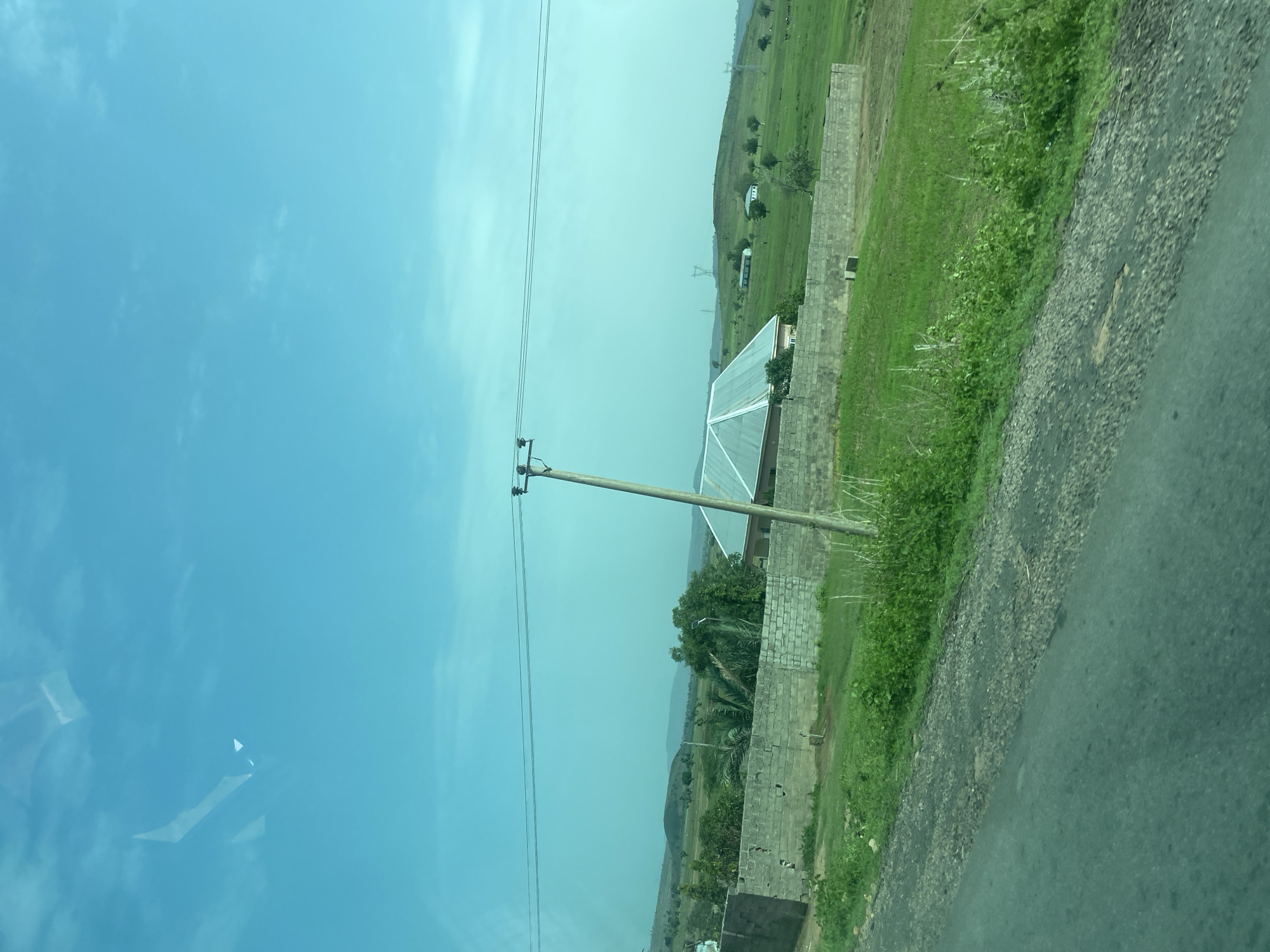 This image depicts a rural landscape, likely in the vicinity of Chum, Nigeria, under a bright, hazy sky. A paved road with visible cracks and gravel runs along the bottom and left side of the frame, suggesting a route through the area. Adjacent to the road is a strip of overgrown green vegetation.
Further into the scene, a low, brick wall encloses a property. Behind the wall, a house with a metallic, gabled roof is visible. The house is surrounded by lush green grass and some scattered trees, including a palm tree. A concrete utility pole with wires stretches across the upper portion of the image.
In the background, rolling green hills rise, dotted with trees and some distant structures, possibly other houses and power transmission towers. The overall impression is of a serene, sparsely populated rural environment. There are no people or animals clearly discernible in the image.