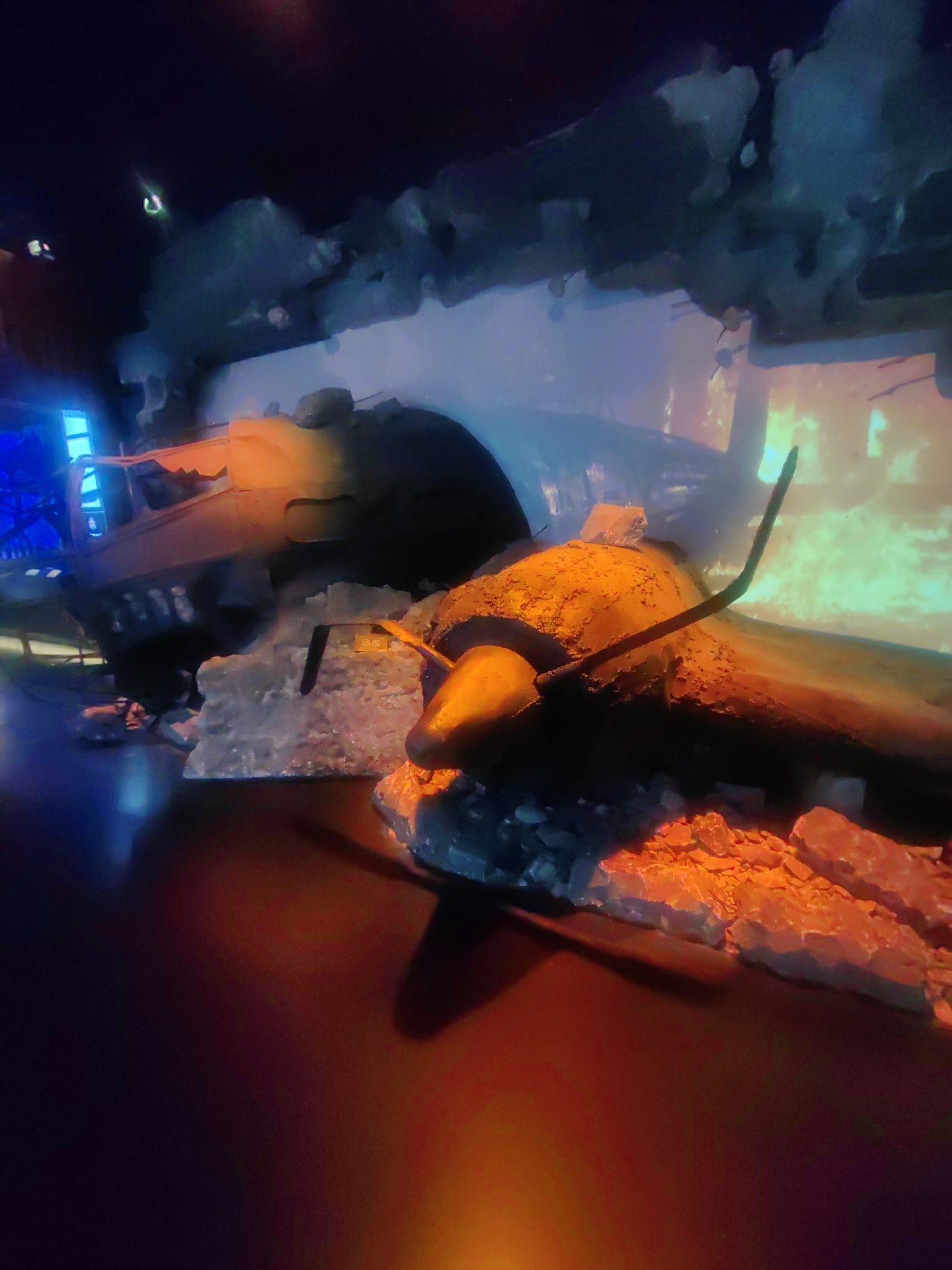 The image depicts a dimly lit, theatrical diorama featuring what appears to be a crashed or damaged aircraft prop. The foreground is dominated by a dark, reflective surface, likely a floor, with patches of orange and red light emanating from below and to the right. Scattered across this surface are chunks of what resembles ice or rough-hewn rock, some illuminated by the orange light.
The central subject is a rusty, orange-brown object that might be a section of the aircraft's fuselage or a large engine component, with a bent, dark metal rod protruding from it, resembling a broken propeller or antenna. This object rests on a base of the same icy or rocky material.
In the background, a larger structure, possibly another part of the aircraft or a backdrop, is visible, rendered in muted blues and grays, with a luminous quality that suggests internal lighting or projection. The overall scene evokes an impression of a crash site or a display in an exhibition related to aviation or a disaster scenario. There is no indication of the location being Zelenaya Roshcha, Russia, as the image provides no contextual clues to support this.
