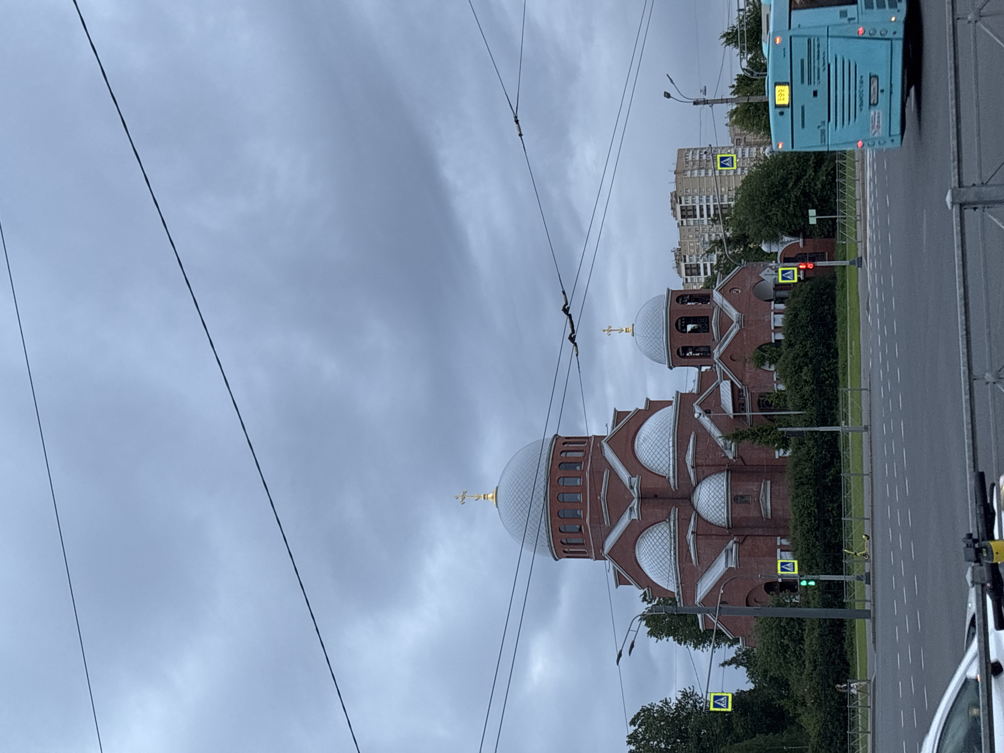 The image depicts an urban scene in Unknown, Unknown, dominated by a red brick church with multiple silver domes topped with golden crosses. The sky is overcast with gray clouds. Power lines stretch across the foreground, obscuring the view. To the right of the church is a road with pedestrian crosswalks and traffic lights. A blue bus is partially visible at the top right, and a white car is seen at the bottom right. The traffic lights show a green signal, indicating permission to proceed. Trees and bushes are present along the roadside. Buildings can be seen in the background behind the trees.