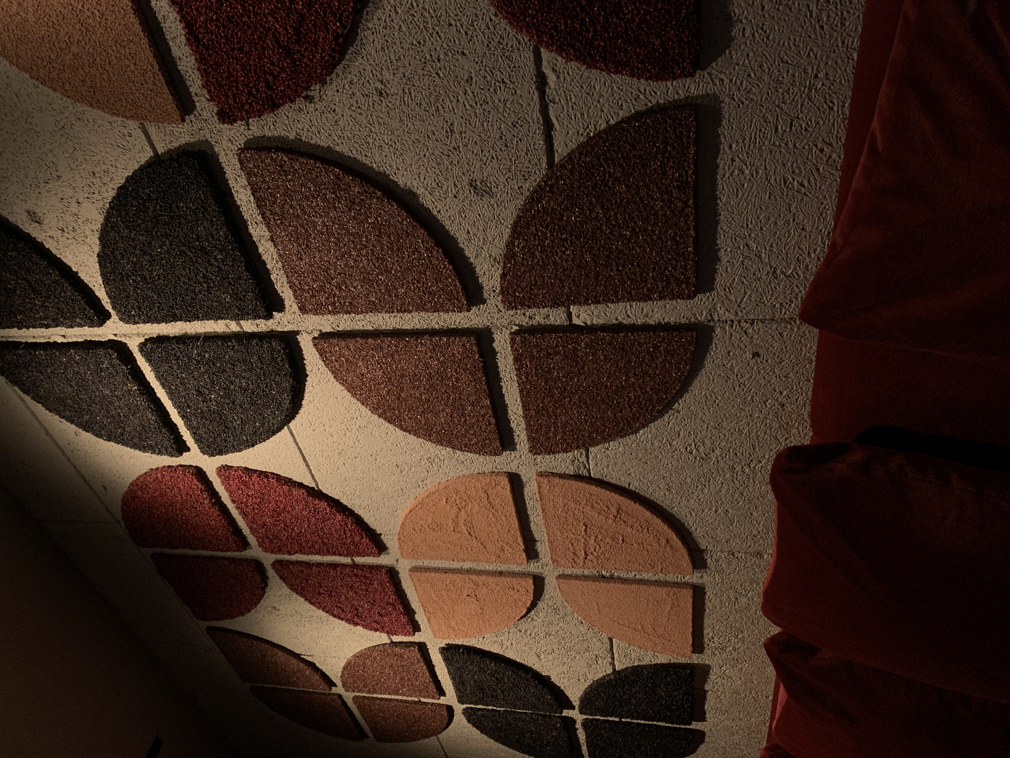 The image presents an angled, close-up view of an interior surface, likely a wall or ceiling, adorned with decorative and potentially acoustic panels. The background features a light gray, fibrous texture, characteristic of wood wool or a similar sound-absorbing material.
Arranged across this surface are numerous geometric shapes, primarily semi-circles and quarter-circles, forming abstract patterns. These shapes exhibit a coarse, granular texture and come in various earthy and muted colors, including dark brown, reddish-brown, black, and beige. They are often grouped to create larger circular motifs or intricate tessellations. Strong, directional lighting from the upper left casts prominent shadows beneath these panels, highlighting their raised, three-dimensional nature and adding depth to the composition.
On the far right side of the frame, a section of draped, deep reddish-brown or dark red fabric is visible. Its soft, folded appearance suggests it could be part of a curtain, upholstery, or a decorative textile element.
No people are present, and no specific activity is occurring. There is no visible text. The lighting conditions suggest an indoor environment, likely illuminated by artificial sources, contributing to the dramatic interplay of light and shadow on the textured surfaces. The overall impression is of a designed space focusing on texture, color, and geometric forms.