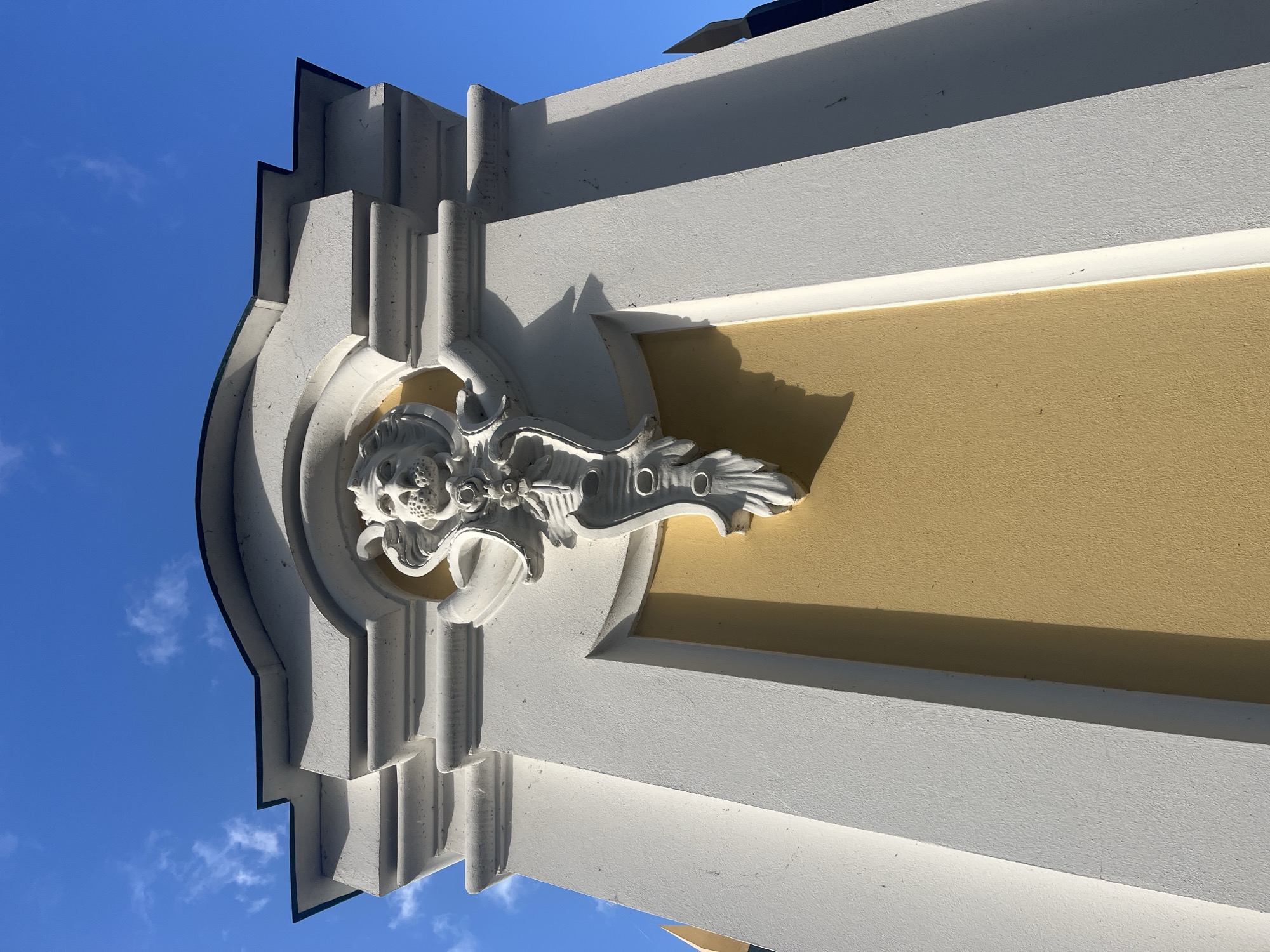 The image shows a close-up, low-angle view of the facade of a building, likely in Peterhof, Russia, due to its architectural style. The building features ornate white stucco ornamentation against a textured yellow wall. Prominently displayed is a decorative relief of a lion's head with a flowing mane, rendered in white stucco. Below the lion's head, a vertical decorative element with circular and leaf-like motifs hangs down. The building's architectural elements include layered white moldings and cornices, casting shadows that highlight their three-dimensional forms. The upper portion of the image reveals a clear, bright blue sky with a few wispy white clouds. The overall scene is well-lit, suggesting daylight.