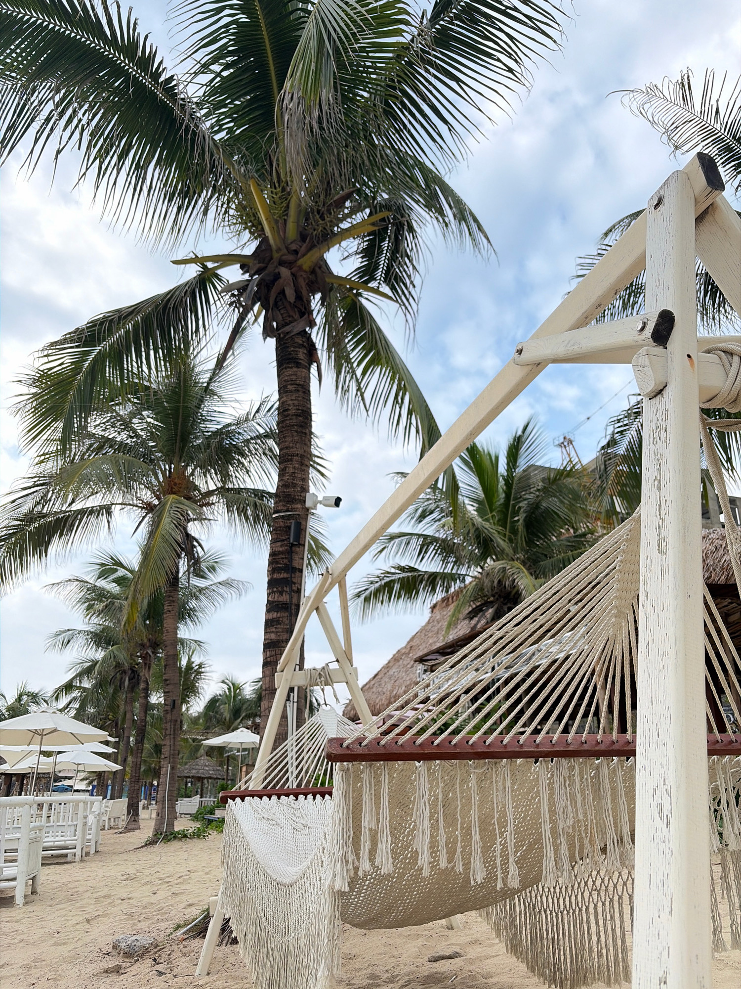 A white wooden hammock frame stands on a sandy beach in Đà Nẵng, Vietnam, with a white macrame hammock suspended from it. The hammock features intricate knotting and fringe details. In the background, several palm trees with lush green fronds sway gently against a cloudy sky. A white security camera is mounted on one of the palm trees. Further back, there are rows of white tables and chairs, some partially covered by white umbrellas, suggesting a resort or beachside dining area. A thatched-roof structure is visible behind the hammock. The scene conveys a relaxed tropical atmosphere.