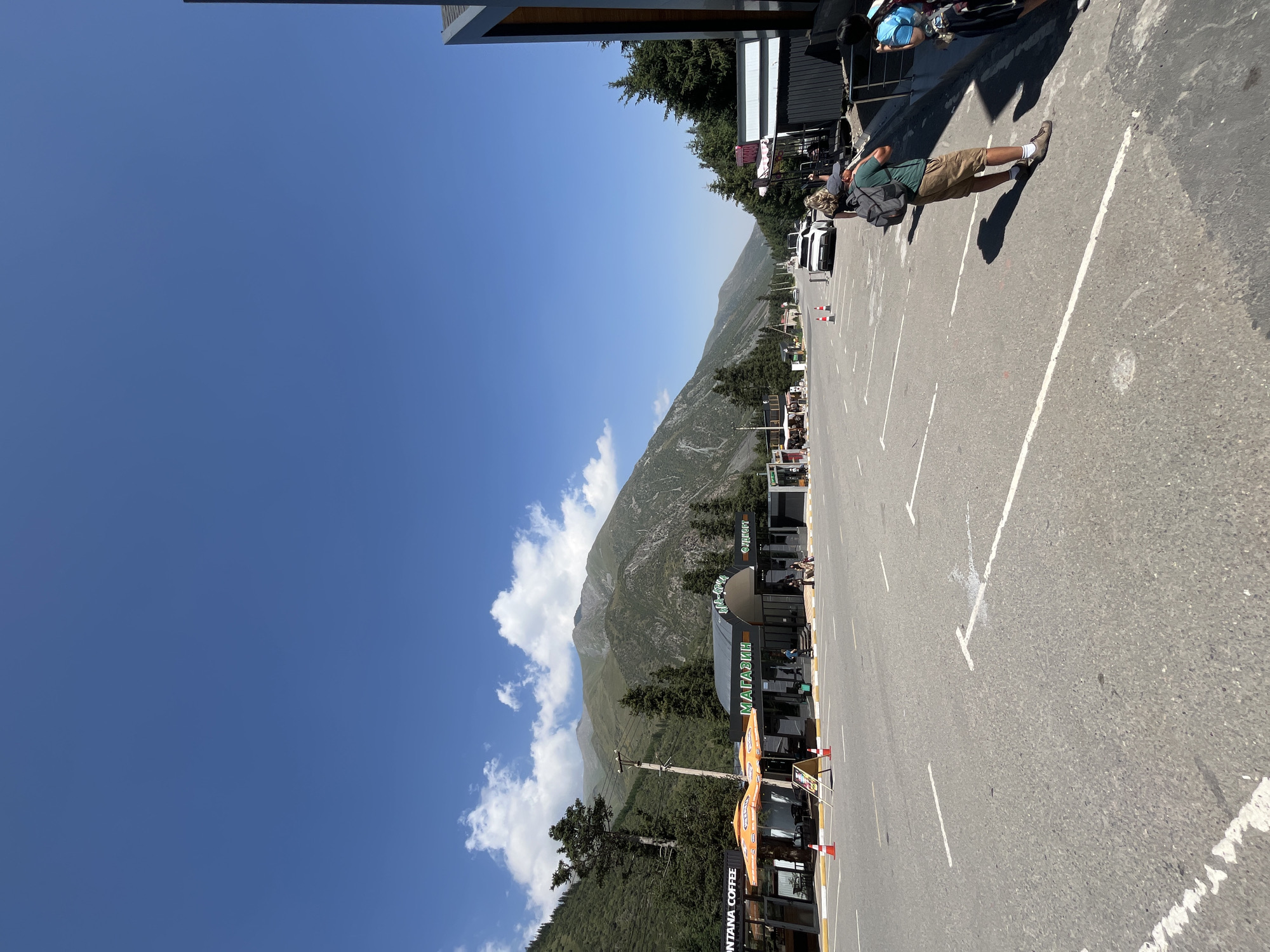 This is a wide-angle, eye-level photograph taken outdoors on a sunny day, looking across a paved road towards a mountainous landscape. The road appears to be a parking area with white painted lines indicating parking spaces. In the mid-ground, there are several commercial establishments with awnings and signs. To the left, there is a building labeled "MONTANA COFFEE" with orange umbrellas outside. Next to it is a building with green lettering that appears to say "МАГАЗИН" (MAĞAZIN, meaning "shop" in Russian). Further down the road, there are more shops and some vehicles, including a white car.
On the right side of the road, a man wearing a green t-shirt, khaki shorts, and a backpack is standing with his back to the camera, facing the parked cars. He appears to be looking at his phone. A woman with blonde hair is standing near him, also facing away from the camera. Further to the right, another person is partially visible.
The background features a steep, tree-covered mountain range under a clear blue sky with scattered white clouds. The overall setting suggests a roadside stop or a small commercial area in a mountainous region, possibly a tourist destination. The location is identified as Kashka Suu, Kyrgyzstan.