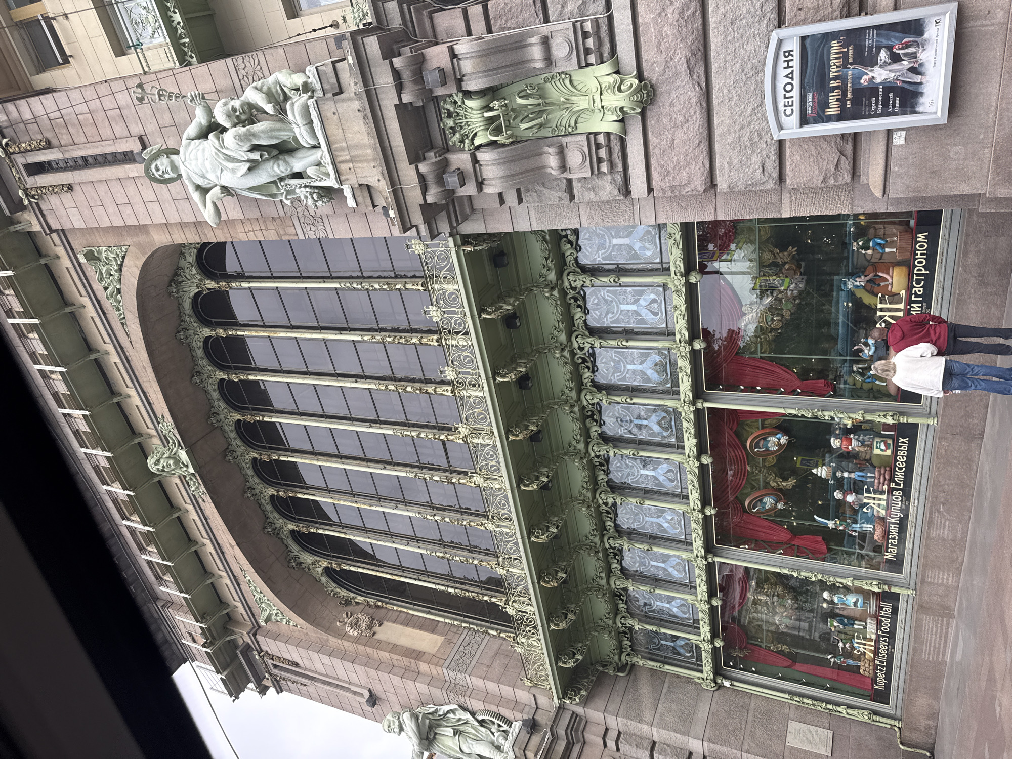 The image captures a partial view of the facade of the Eliseev Emporium in Saint Petersburg, Russia. The architectural details are prominently displayed, with large windows adorned with elaborate wrought ironwork and ornate decorations. Above the windows, there are sculptural elements. The windows display mannequins and food items.
A sign advertising a theatre performance "Night at the Theatre" is visible. Two people are standing in front of the windows. The street scene is typical of the area.