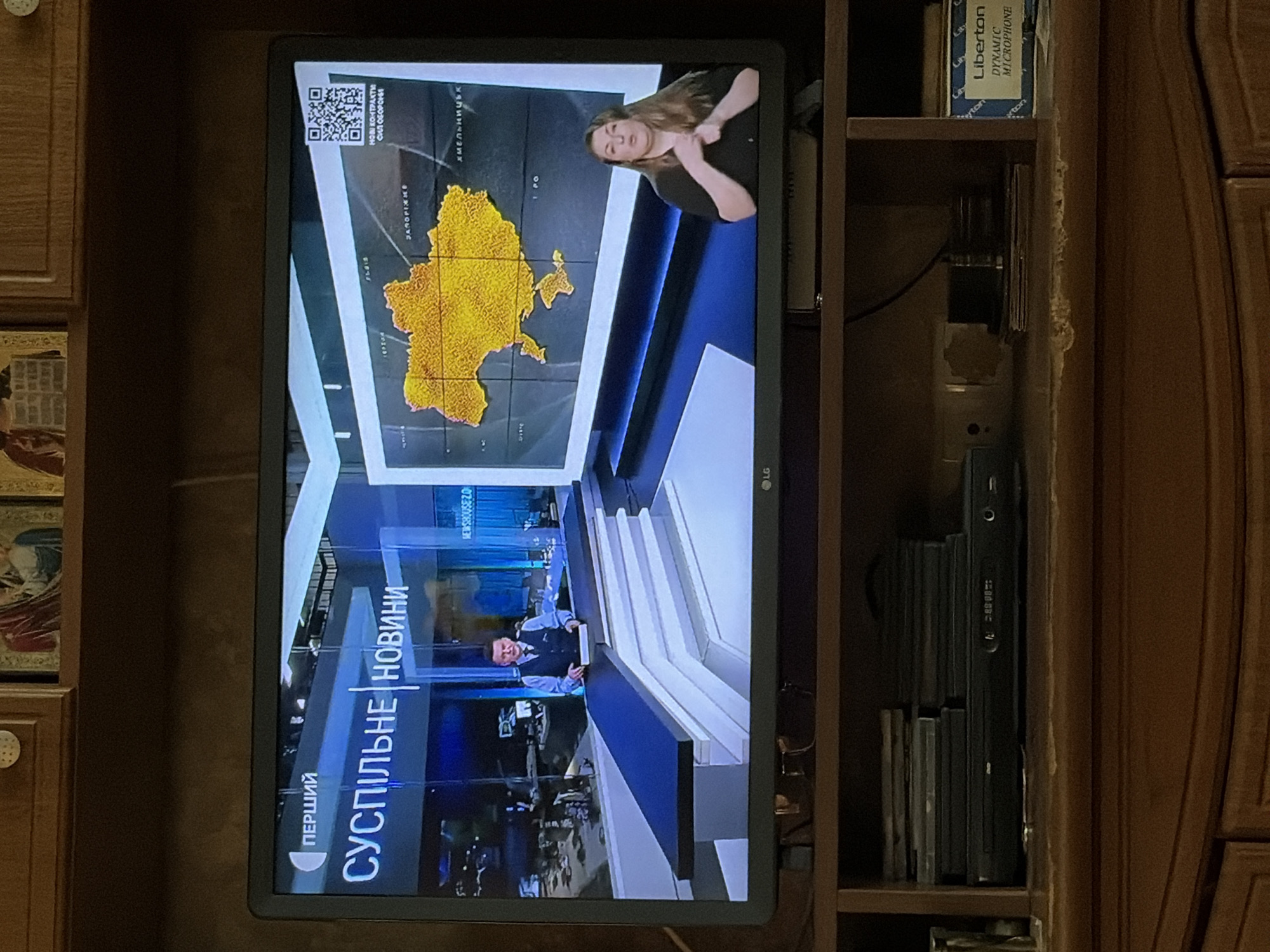The image shows a television screen displaying a news broadcast. On the screen, a male news anchor is standing behind a desk in a studio setting. To the right of the anchor, a map of Ukraine is displayed on a large screen, with a woman performing sign language in front of it. The television is placed on a wooden entertainment center, with stacks of DVDs and a DVD player visible below. A box labeled "Liberton DYNAMIC MICROPHONE" is also on the entertainment center. The overall environment suggests a home setting where a news program is being watched.