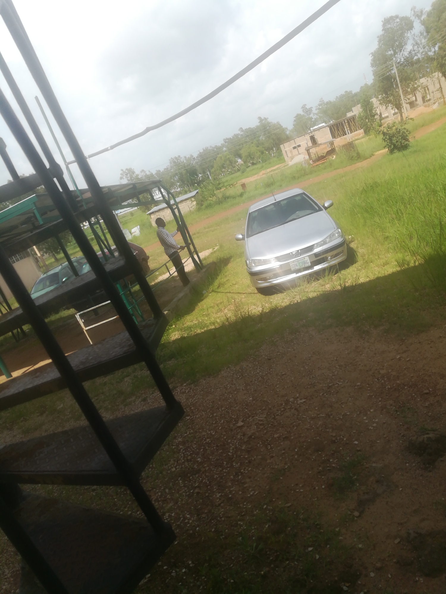 An outdoor, eye-level, medium shot captures a construction site in Bisichi, Nigeria. In the foreground, a dark metal staircase with worn treads is visible, leading upwards and out of frame to the left. To the right of the staircase, a silver sedan is parked on a grassy area. The car has its headlights on and a license plate that reads "GBK 739HG". Beyond the car, a man in a checkered shirt and dark pants stands near a small, partially constructed building with wooden supports. Further in the background, more unfinished buildings with scaffolding are visible, surrounded by lush green trees. The sky is overcast with diffused light.