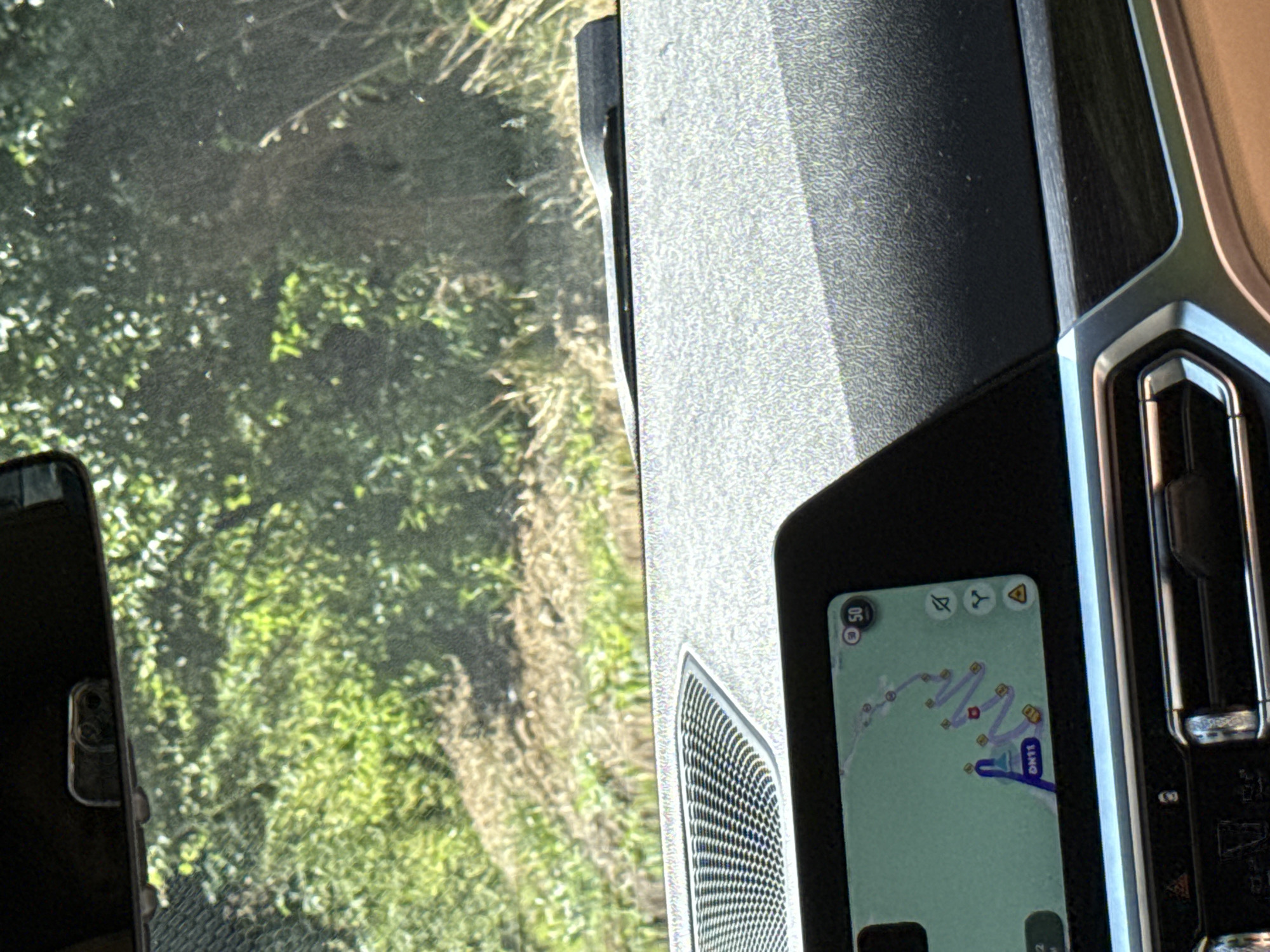 The image shows the interior of a car, looking out through the windshield. The dashboard is visible in the foreground, with a navigation screen displaying a winding route. The windshield reflects the green foliage of trees and bushes, indicating an outdoor, likely rural, environment. The scene appears to be captured during daylight. The location is identified as Oituz, Romania.