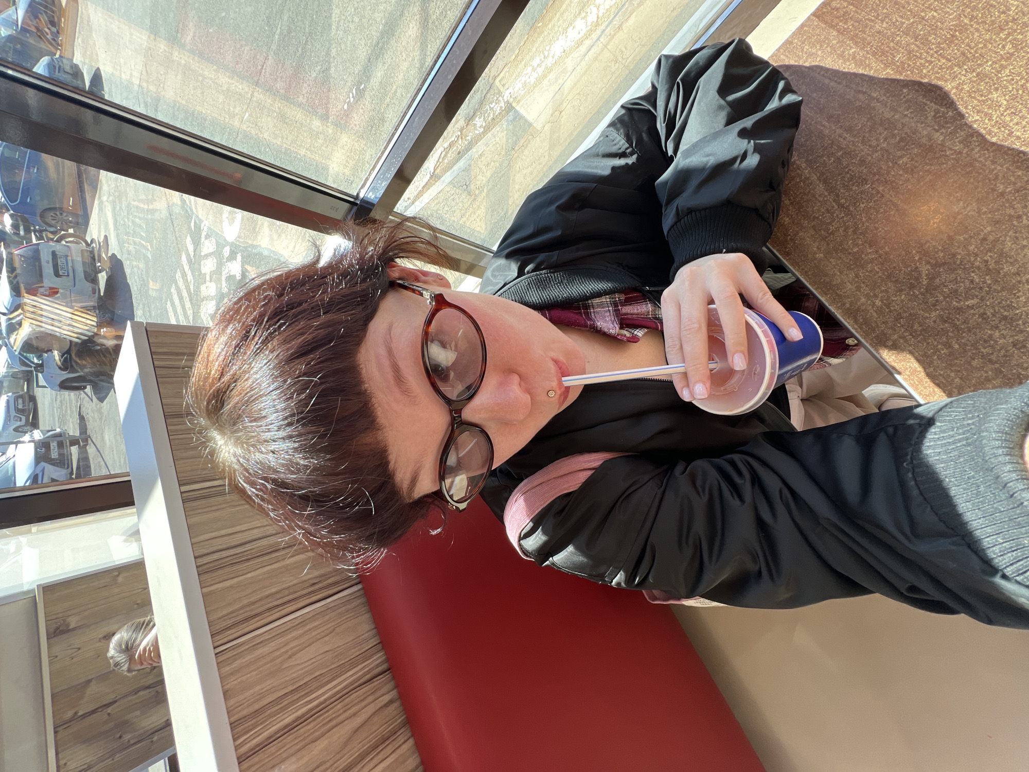 A person is seated at a brown-speckled table next to a large window, likely in a cafe or diner setting. The image is taken from a slightly upward angle, showing the person from the chest up, angled to the right. They have dark brown hair, which catches the bright sunlight streaming in from the window. They are wearing a black bomber jacket over a plaid shirt with dark red and black patterns. Round, tortoiseshell-framed glasses are on their face, and a small stud piercing is visible on their nose. The person is holding a clear plastic cup with a white straw in their right hand, appearing to drink from it. Their fingernails are painted a light, neutral pink or nude color.
Outside the window, a sunny street scene is visible with parked cars. The rear of a light-colored car is visible, bearing a license plate that partially reads "K 265 AT". The bright sunlight casts distinct shadows on the table, indicating it is daytime. The interior features a red booth-style seat. A faint reflection of another person is visible in a lower reflective surface near the table. The overall scene suggests a relaxed moment, enjoying a drink in a well-lit establishment.