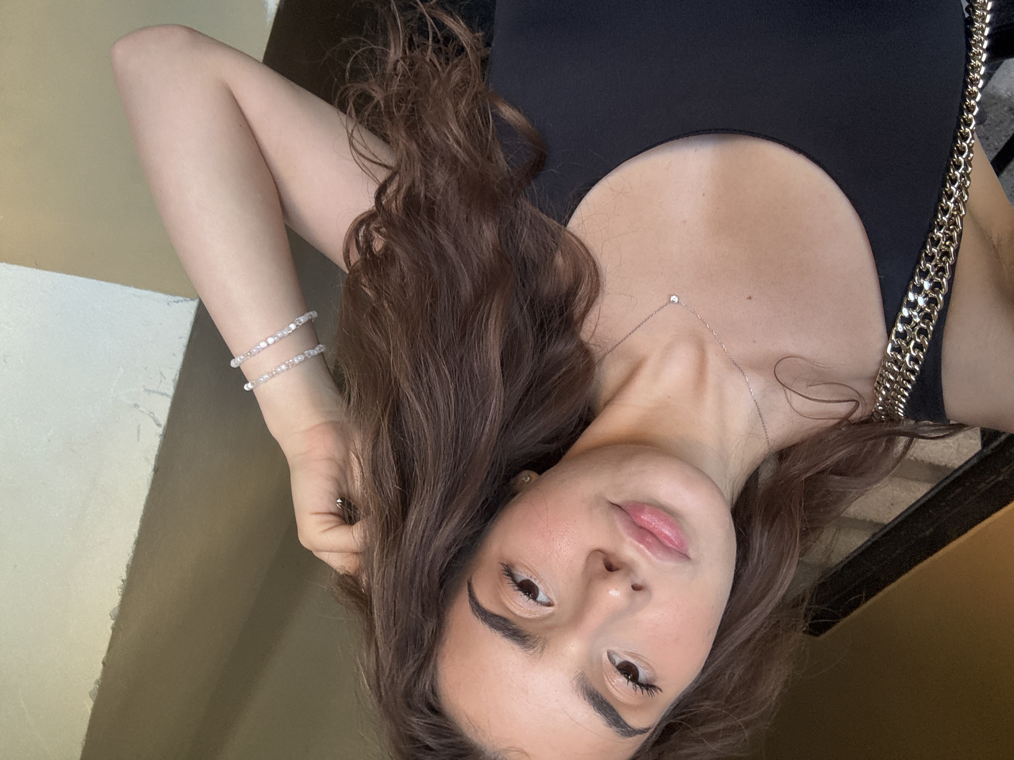 A young woman with long, dark, wavy hair takes a selfie. She is wearing a black tank top and a delicate silver necklace with a small diamond pendant. She also wears two beaded bracelets on her right wrist. The background appears to be an indoor staircase with a wooden railing and a wall with a warm, earthy tone. The lighting suggests it is daytime. There is no discernible connection to the city of Shumen, Bulgaria in the image.