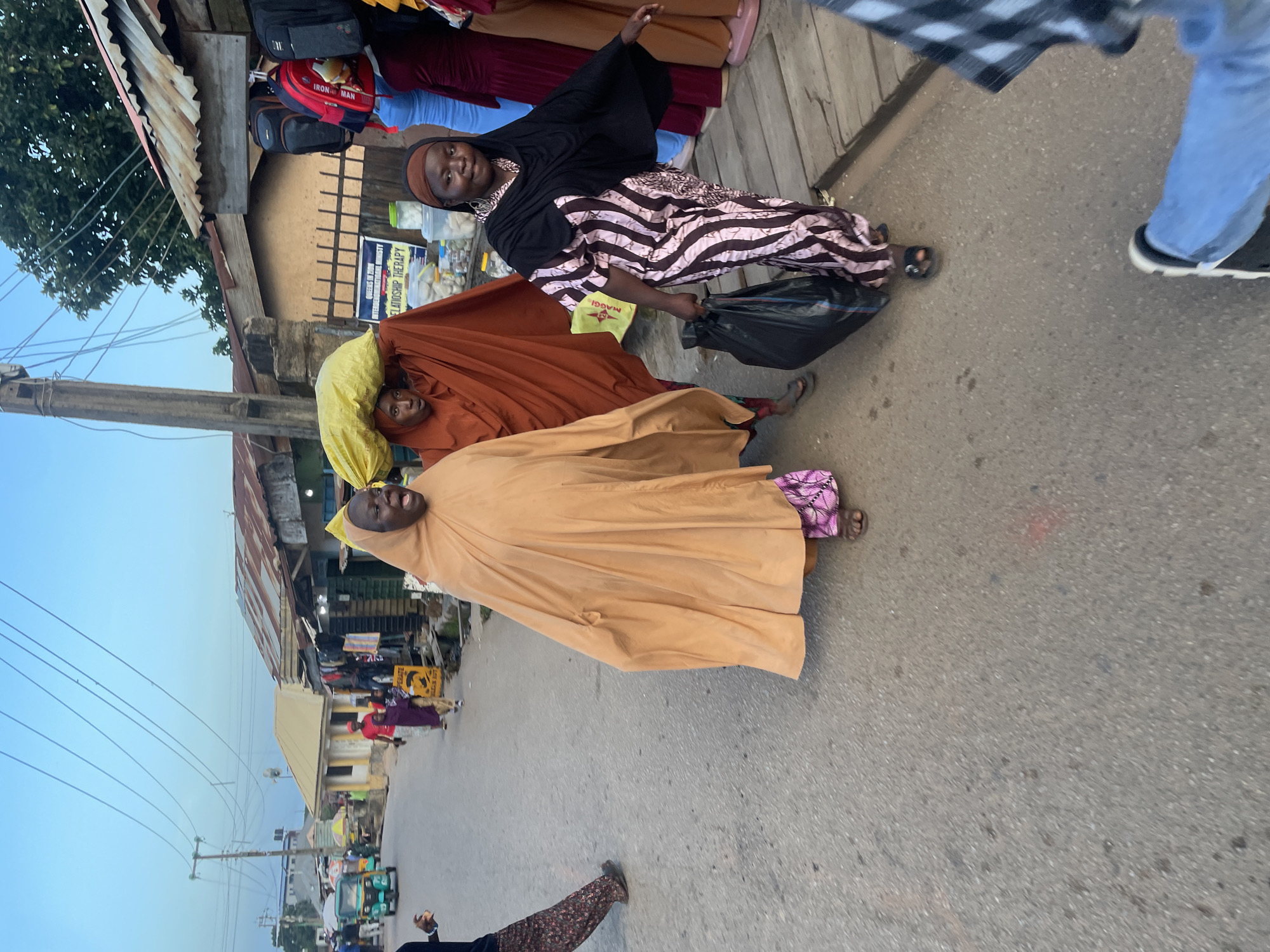 In Jos, Nigeria, on a street with a paved surface, three women are walking. The woman on the left is wearing a flowing, light brown garment and a pink and purple patterned skirt. She appears to be speaking or shouting. To her right, a woman wearing a reddish-brown garment is carrying a large, bright yellow bag on her head. She is looking ahead. The woman on the right is dressed in a black hijab and a pink and white striped dress. She is carrying a black plastic bag in her left hand and looking towards the viewer. The background shows a street lined with shops with corrugated metal roofs. Power lines are visible overhead, and a utility pole stands to the left. A three-wheeled vehicle, possibly a tuk-tuk, is parked in the distance. The overall impression is of a bustling market area or street scene.