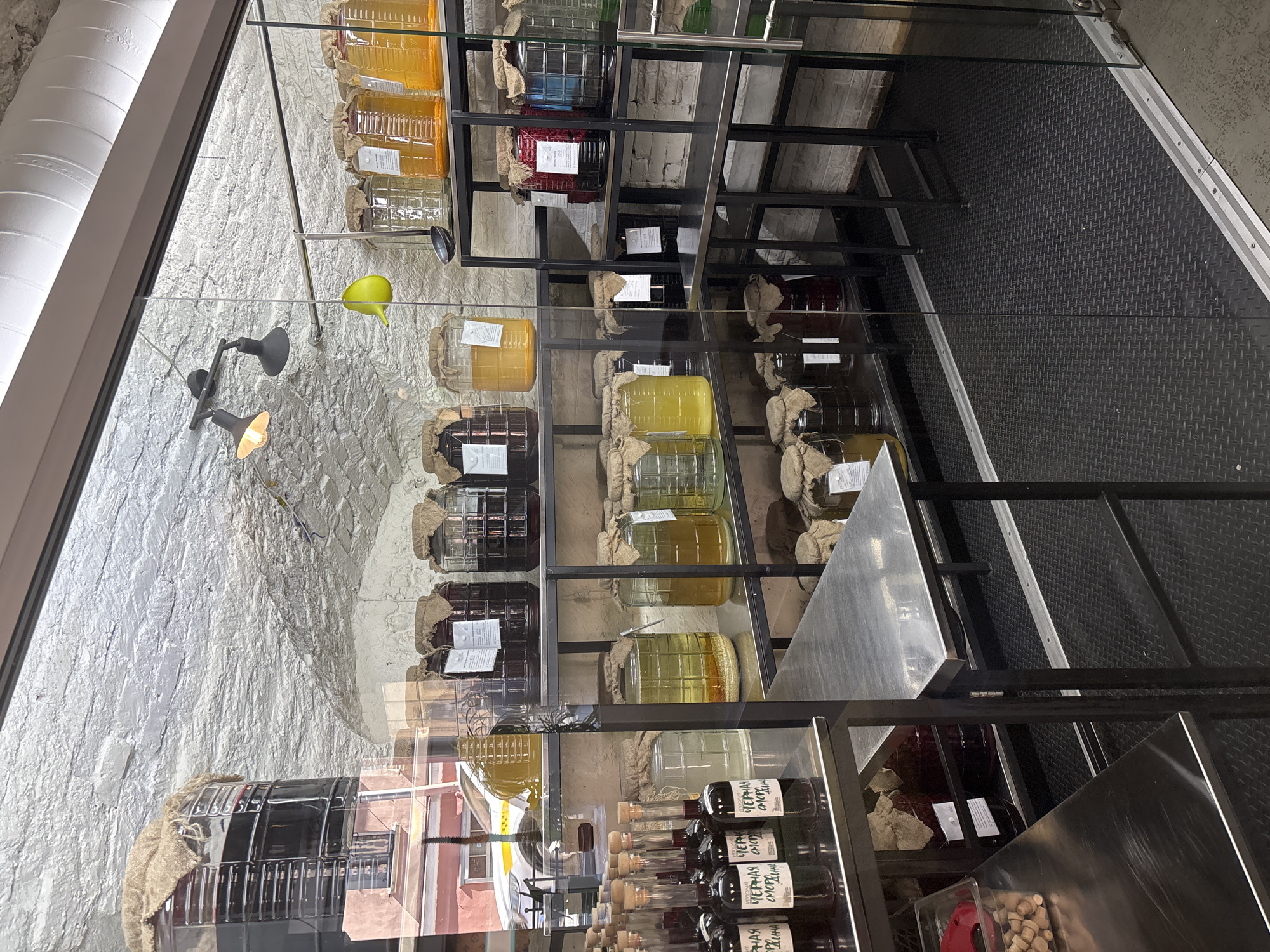 The image displays the interior of a commercial establishment, likely a specialty food or beverage shop, in Saint Petersburg, Russia, viewed through a clear glass partition. The scene is characterized by numerous large glass vessels containing various liquids, set against a rustic-industrial backdrop.
The background features an arched, whitewashed ceiling with a rough texture, and whitewashed brick walls. Two black track lights with warm yellow bulbs illuminate the ceiling, and a bright green, pear-shaped decorative object hangs in the upper-middle. Exposed metal ductwork is visible in the upper right.
Multiple black metal shelving units are arranged throughout the space. These shelves hold numerous large, clear glass jars or demijohns, many of which are covered with a natural, burlap-like fabric secured over their openings. The jars contain liquids of varying colors, including dark reds, amber yellows, and pale or clear liquids; some show sediment at their base. Rectangular white labels are affixed to many jars.
On the left side, a stainless steel countertop holds rows of smaller clear glass bottles filled with dark liquid and stoppered with corks. Labels on these bottles display "ЧЕРНАЯ СМОРОДИНА" (Black Currant in Cyrillic), indicating the product. Below, a clear plastic container holds small wooden corks. In the upper left quadrant, a large, cylindrical stainless steel vessel, also with a burlap cover, is visible.
The floor is covered with black, textured rubber matting, featuring a diamond-plate pattern. Silver metal tracks delineate the bottom edge of the glass partition.
A reflection on the glass partition reveals an exterior street scene, including part of an orange/pink building with windows and a white car, possibly a taxi due to a visible yellow stripe, parked on the street. No people are visible in the image.