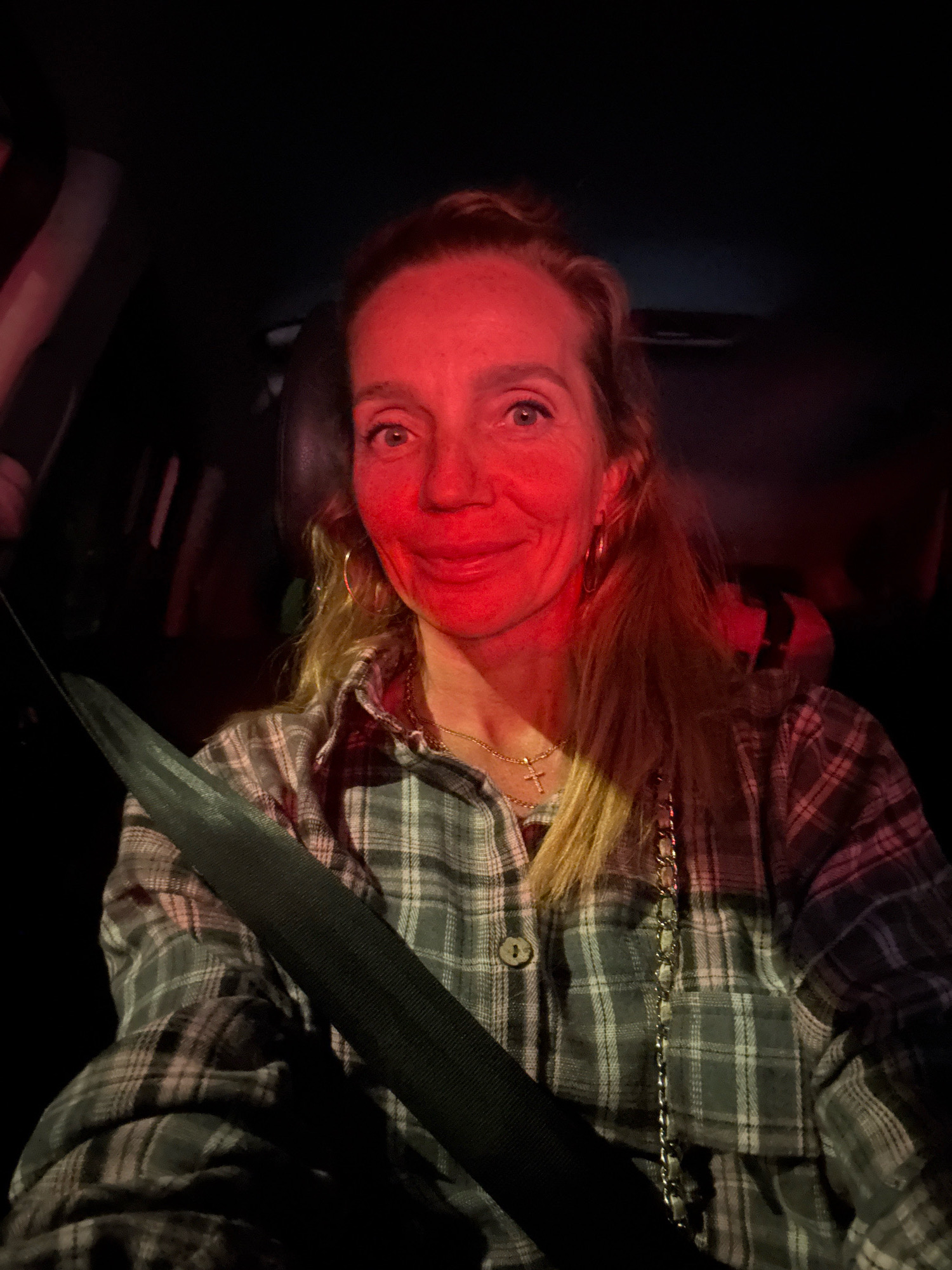 The image shows a woman in a car at night, illuminated by a red light. She is wearing a plaid shirt and a seatbelt. Her face is flushed red from the light, and her eyes are open, looking at the camera. The background is dark and out of focus, suggesting the car is in motion or parked in a dimly lit area. The specific location in Saint Petersburg is not discernible from the image itself.